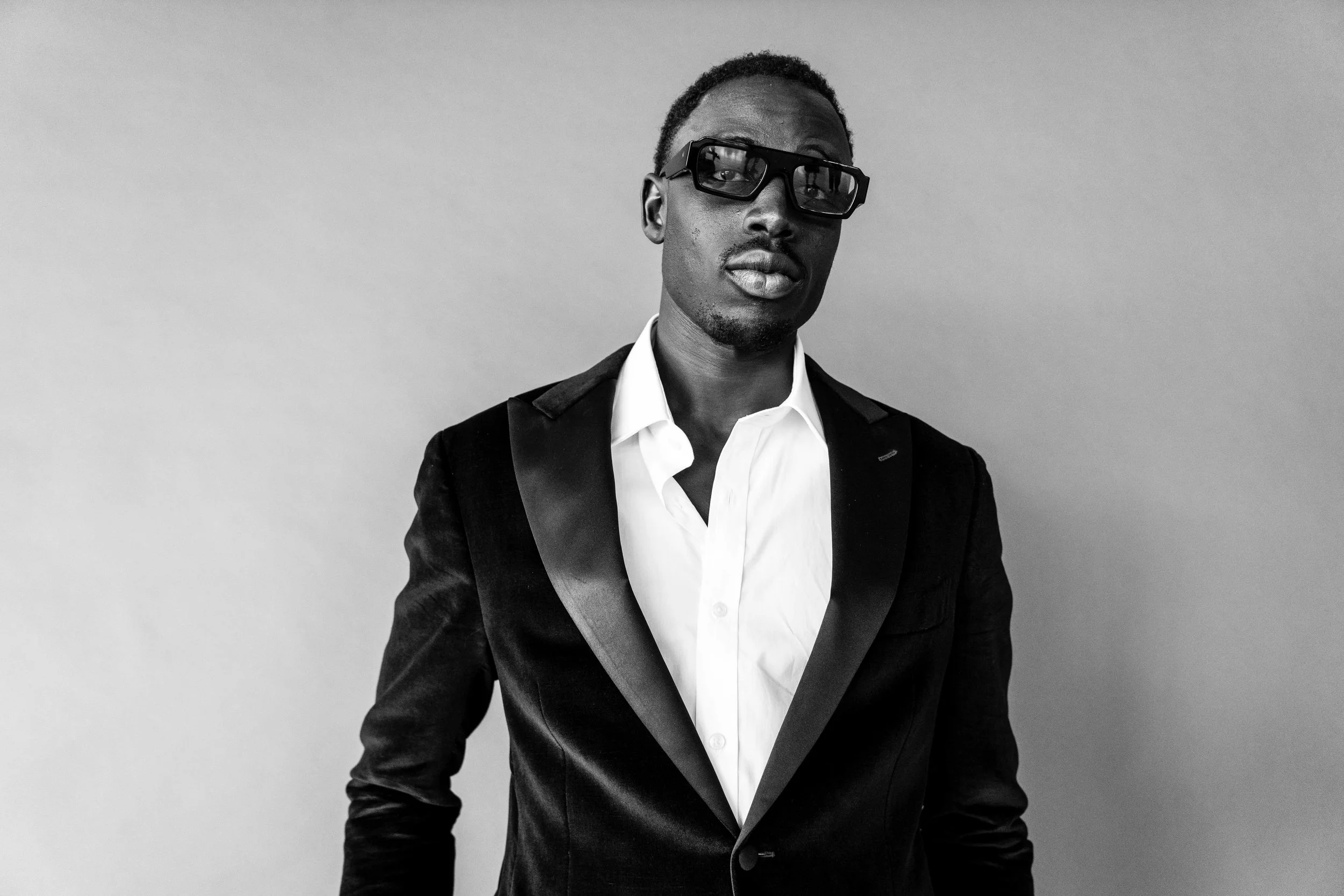 A Black man in a formal suit and white shirt wearing glasses, standing against a plain background.