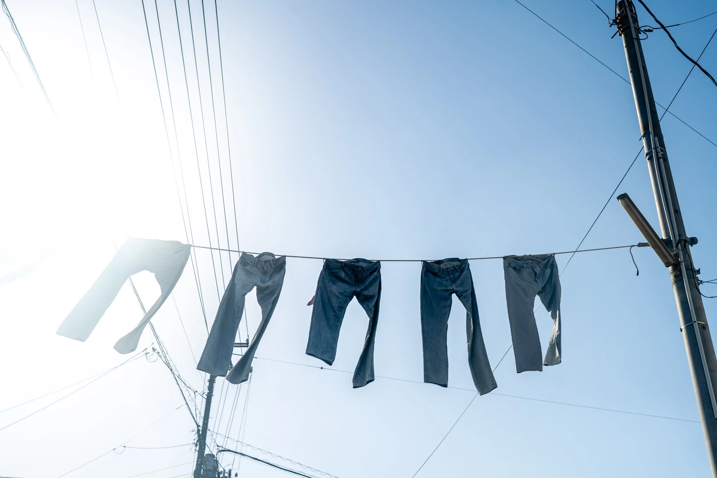Pair of jeans hanging on a clothesline against a clear blue sky with utility poles and wires.