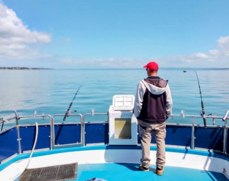 Man fishing on a boat in calm blue water under a partly cloudy sky, wearing a red cap and vest. Out there fishing charters and tours - Westernport Bay