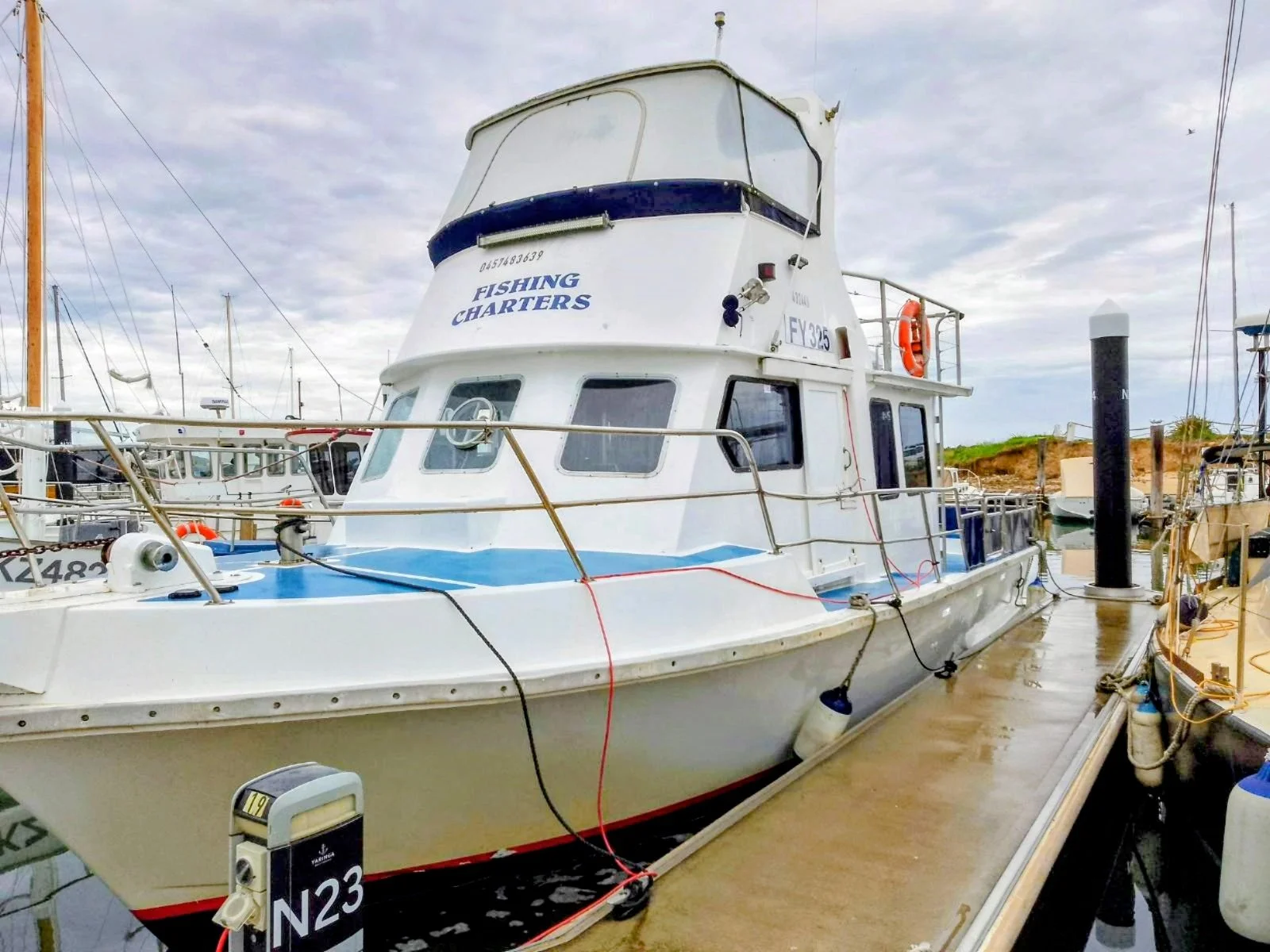 A white fishing boat docked at a marina with other boats, featuring labels 'FISHING CHARTERS', connected to the dock with various ropes and fenders. Out there fishing charters leave from Yaringa Boat Harbour