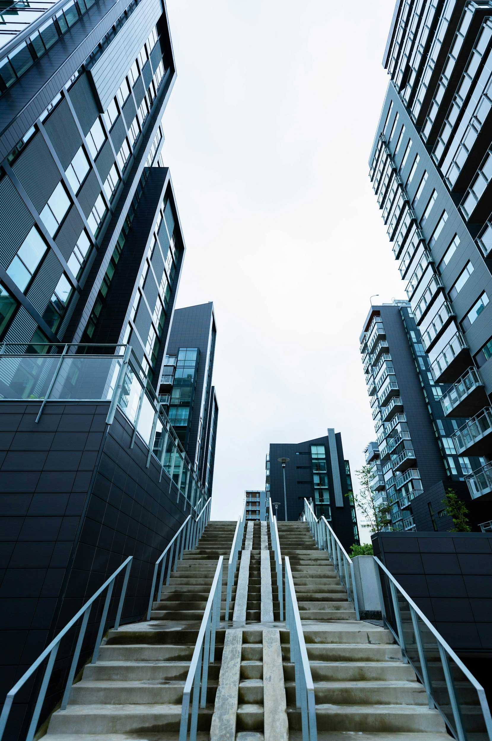 Looking up at a staircase between modern high-rise buildings, with cloudy sky overhead.