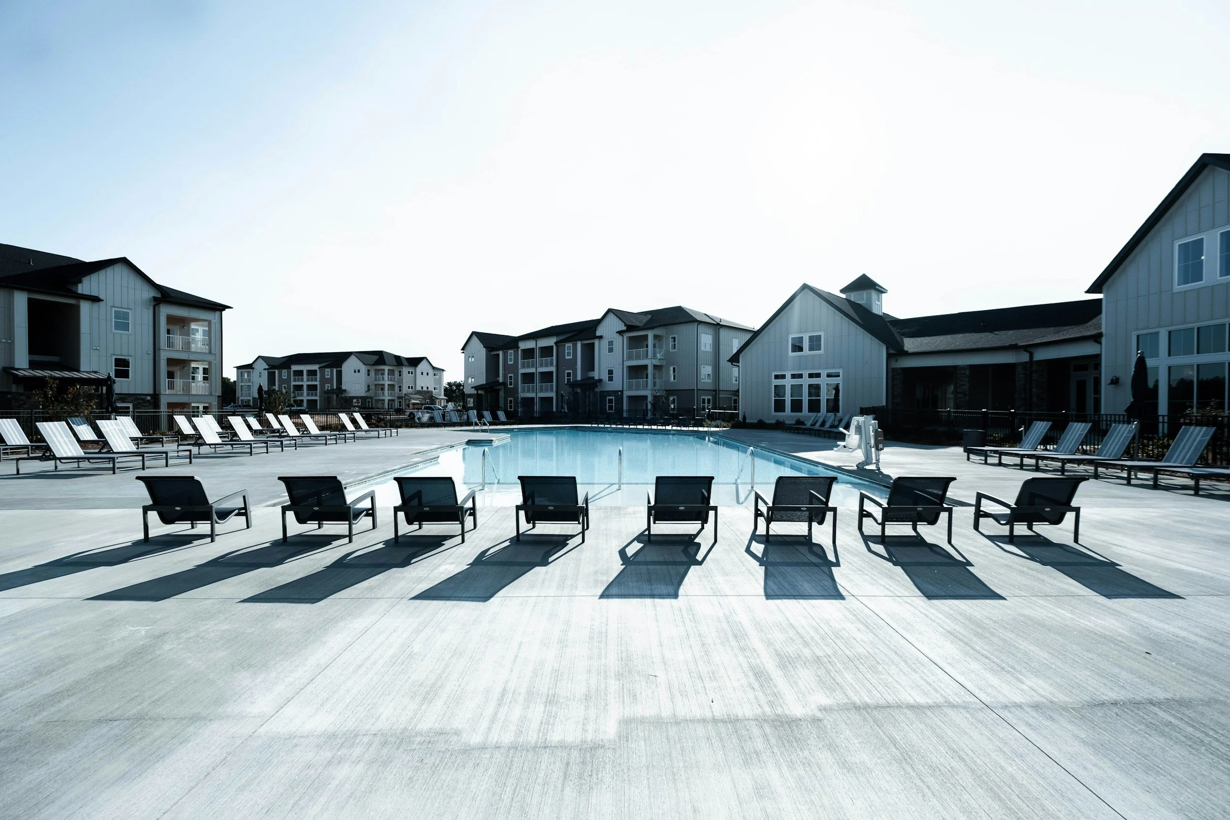 Empty swimming pool surrounded by lounge chairs and residential buildings under bright sky.