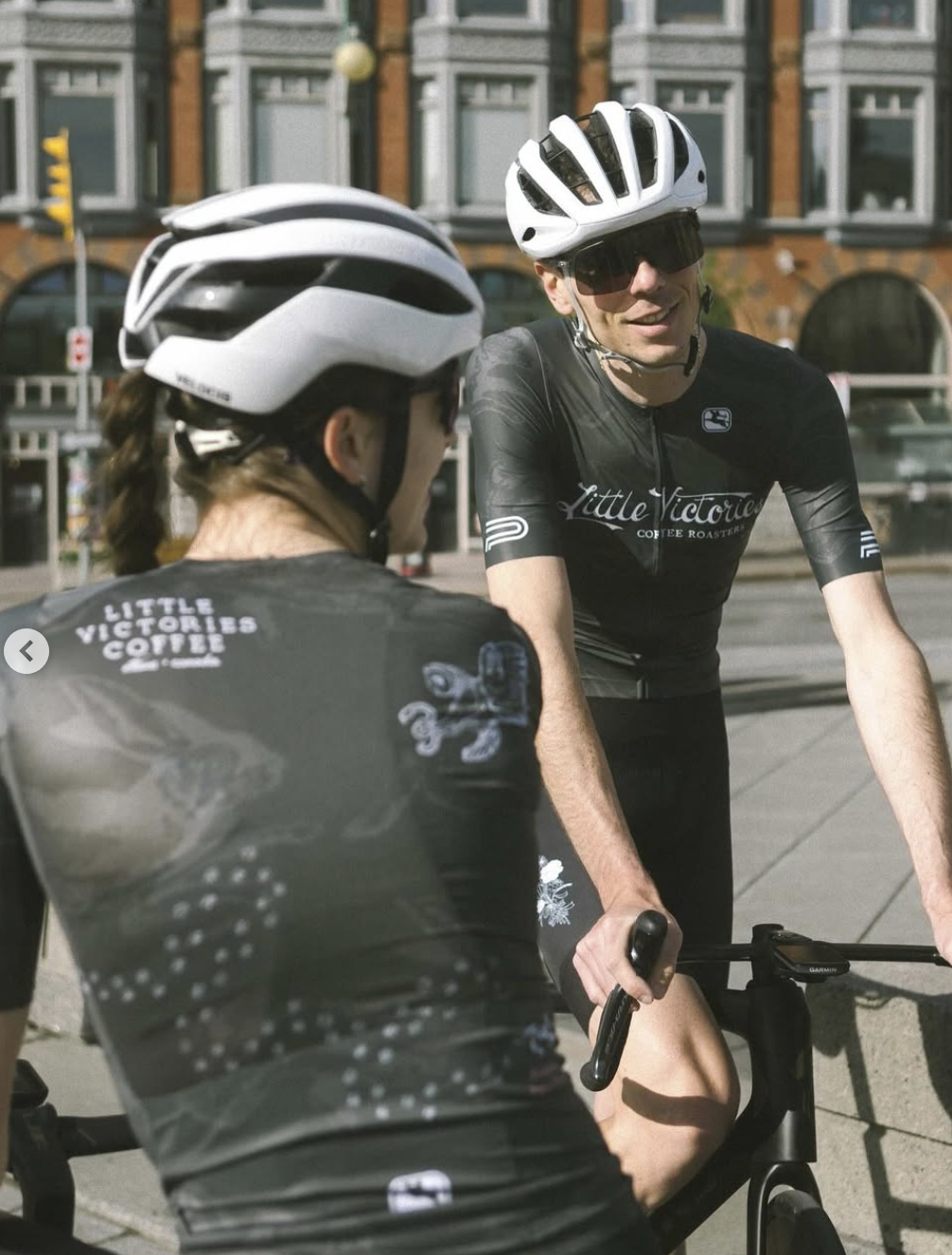 Zwei Radfahrer im Gespräch, beide tragen Helme und schwarze Trikots mit dem Logo "Little Victories Coffee" auf der Straße, im Hintergrund sind historische Gebäude und Straßenverkehrsmittel.