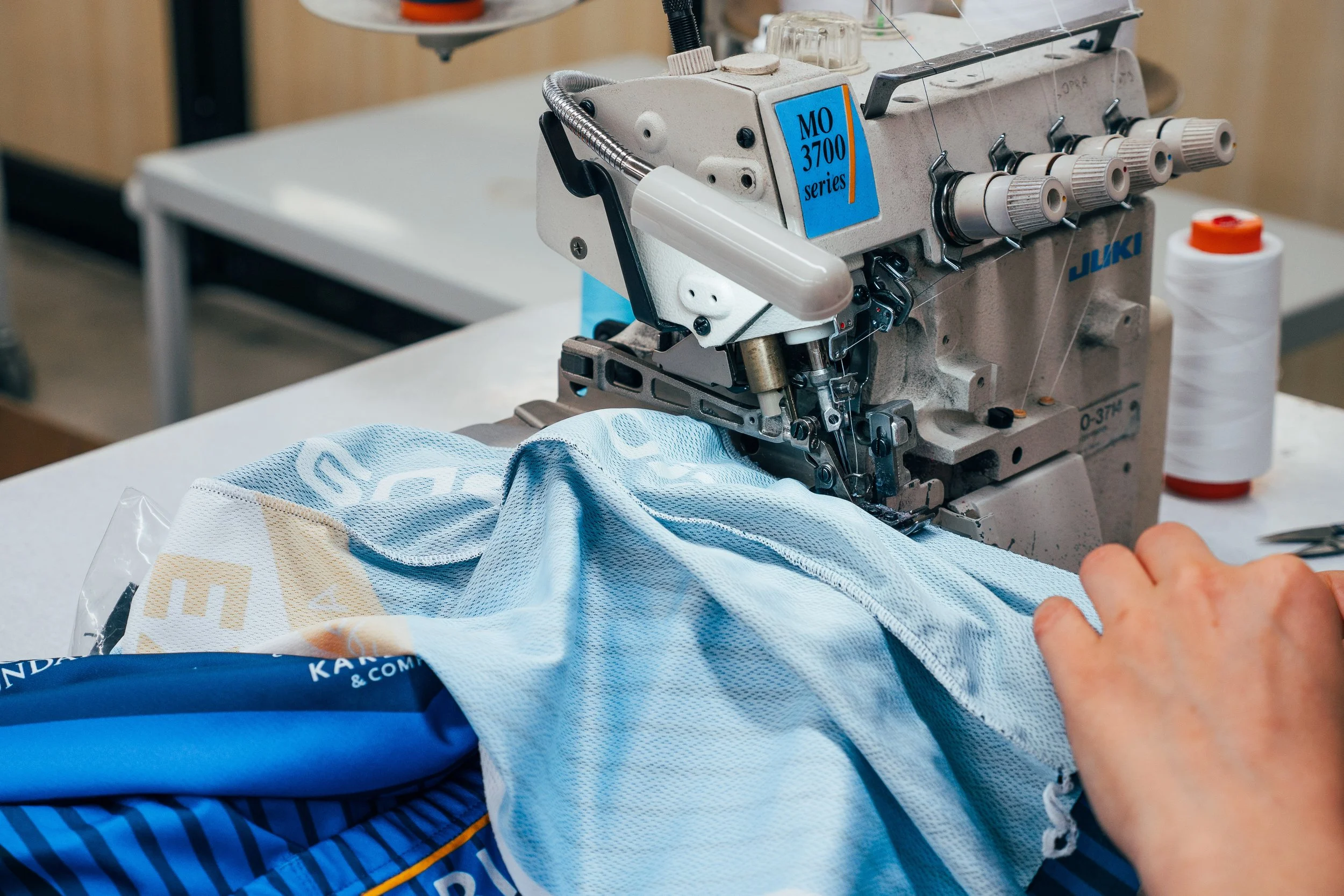 Menschen nähen textiles Material mit einer industriellen Nähmaschine der Marke Juki, auf einem Arbeitstisch, mit Zuführungsschnur im Hintergrund.