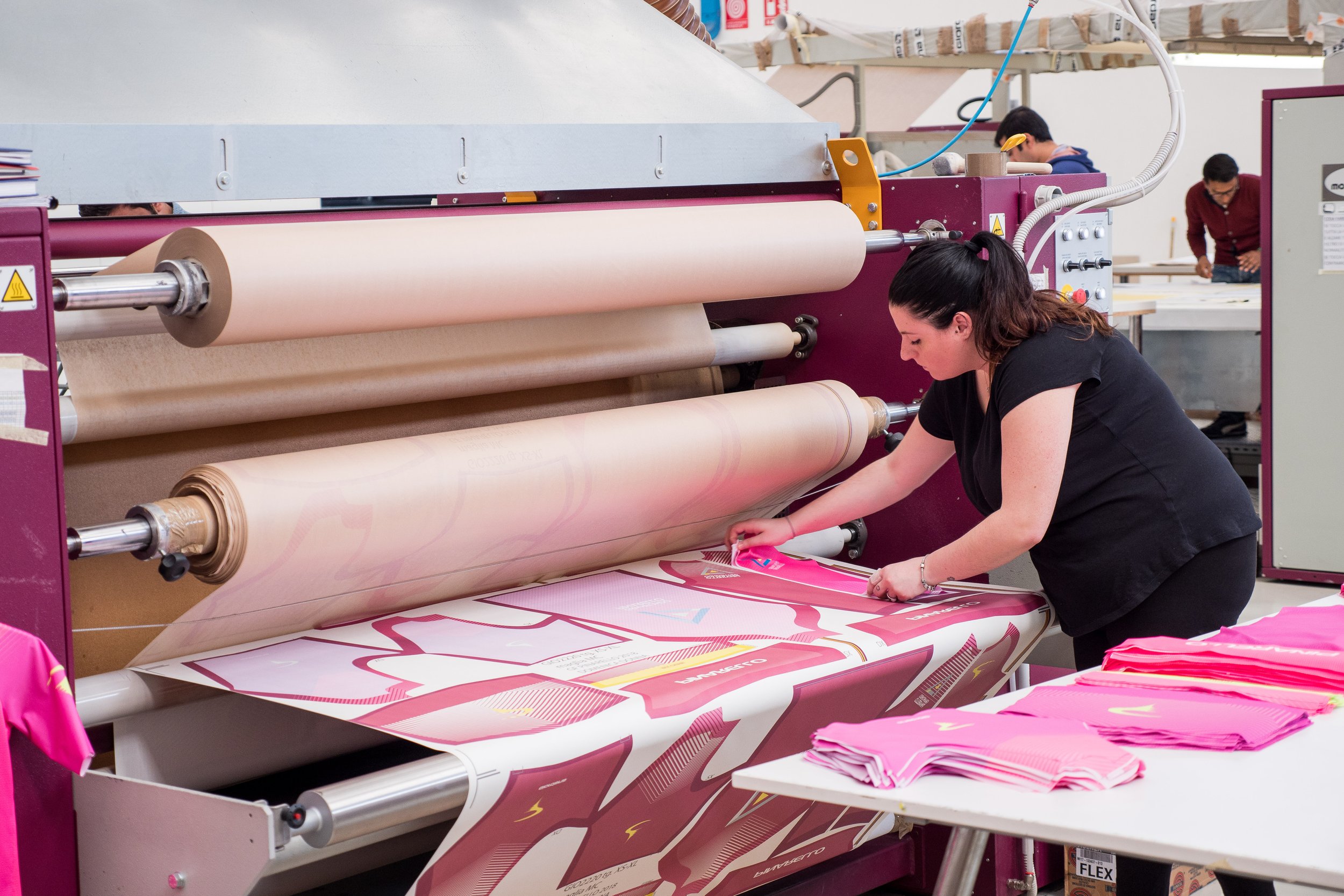 Eine Frau arbeitet an einer großen Druckmaschine, die bunte Papiere druckt. Im Vordergrund liegen fertig gestanzte pinkfarbene Drucke.