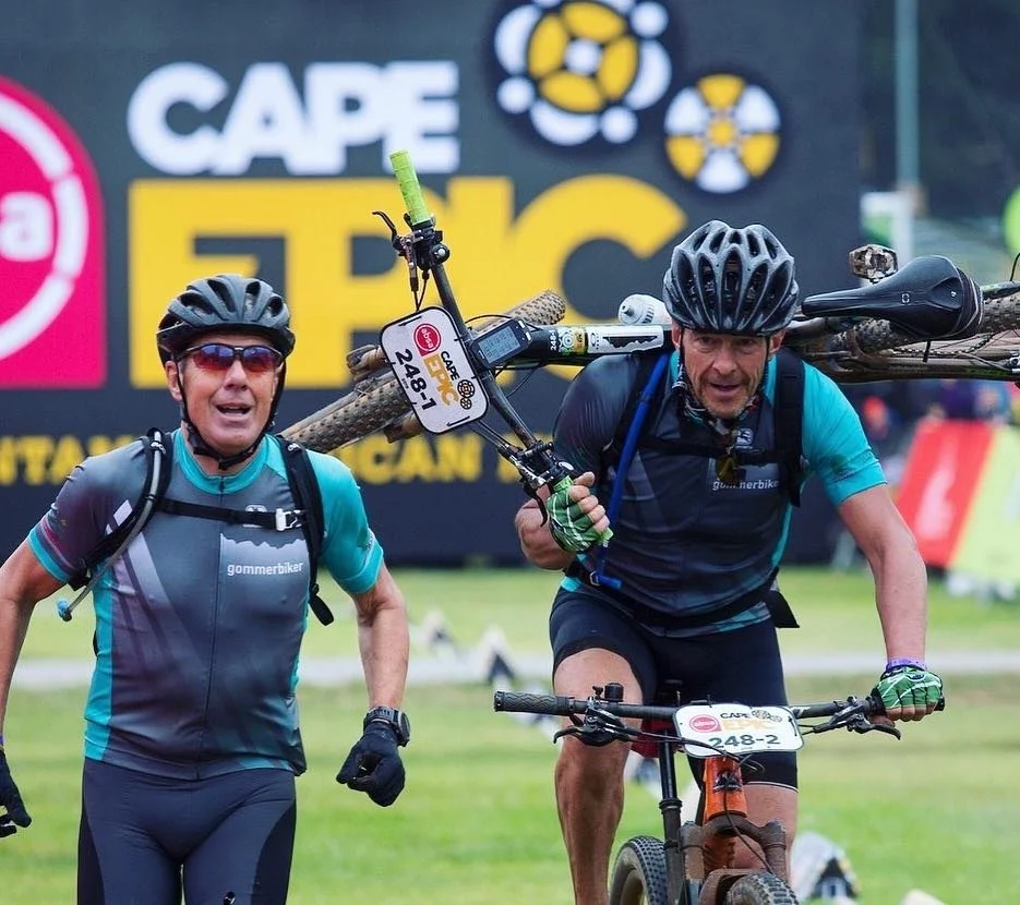 Zwei Mountainbiker, einer trägt eine Holzkonstruktion auf den Schultern während eines Wettkampfs bei Cape Epic. Im Hintergrund ein Cape Epic Logo