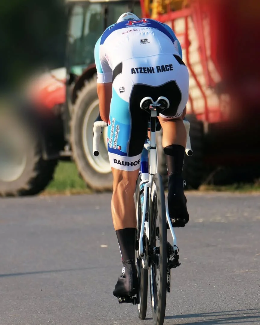 Radfahrer in Rennkleidung auf Fahrrad unterwegs, Rücken zum Betrachter, in Bewegung, auf einer Straße, im Hintergrund landwirtschaftliche Fahrzeuge.