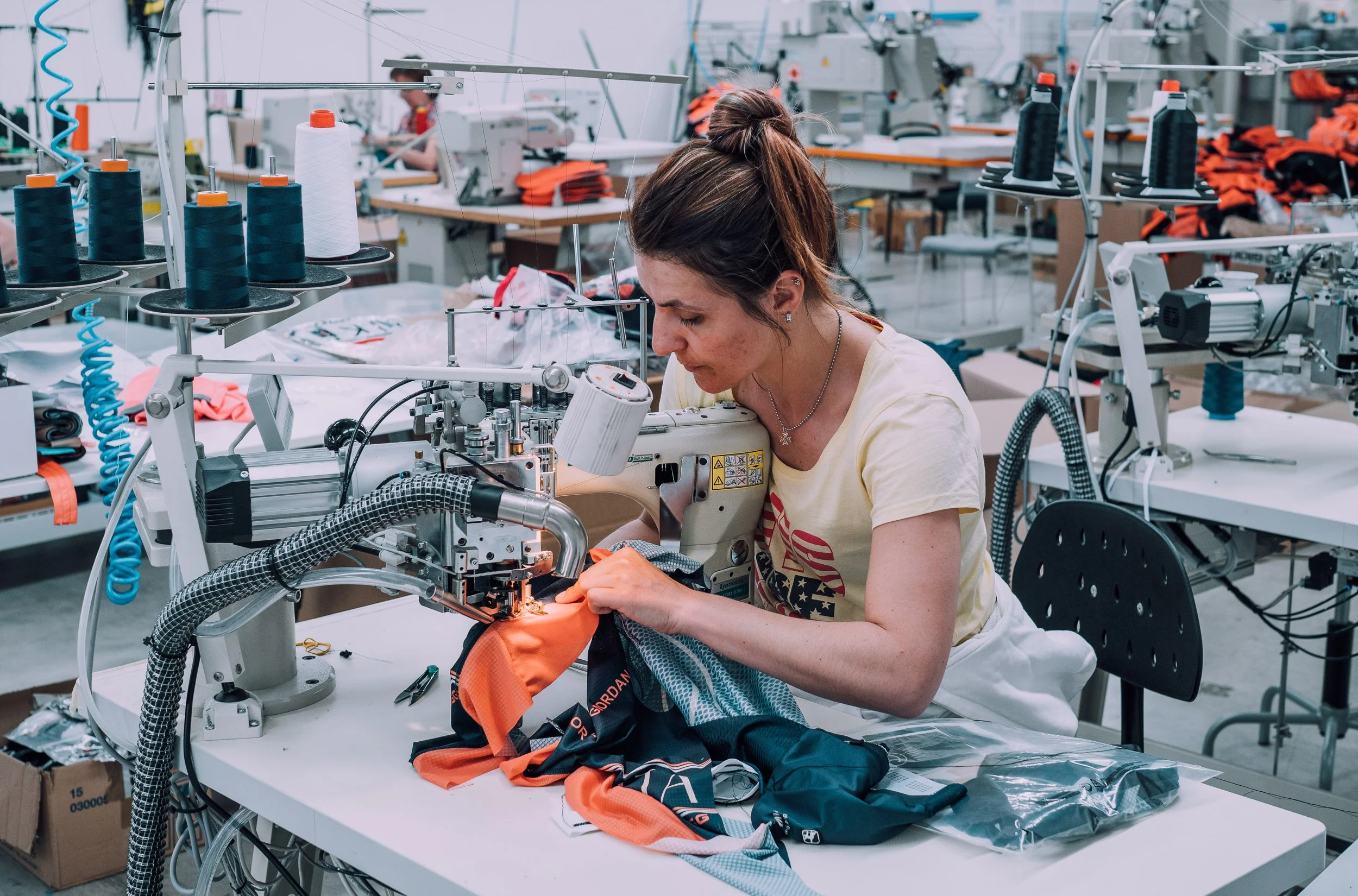 Eine Frau arbeitet in einer Textilfabrik mit einer Industrienähmaschine, näht orangefarbene Stoffe. Im Hintergrund sind weitere Nähmaschinen und Spulen mit Garn.