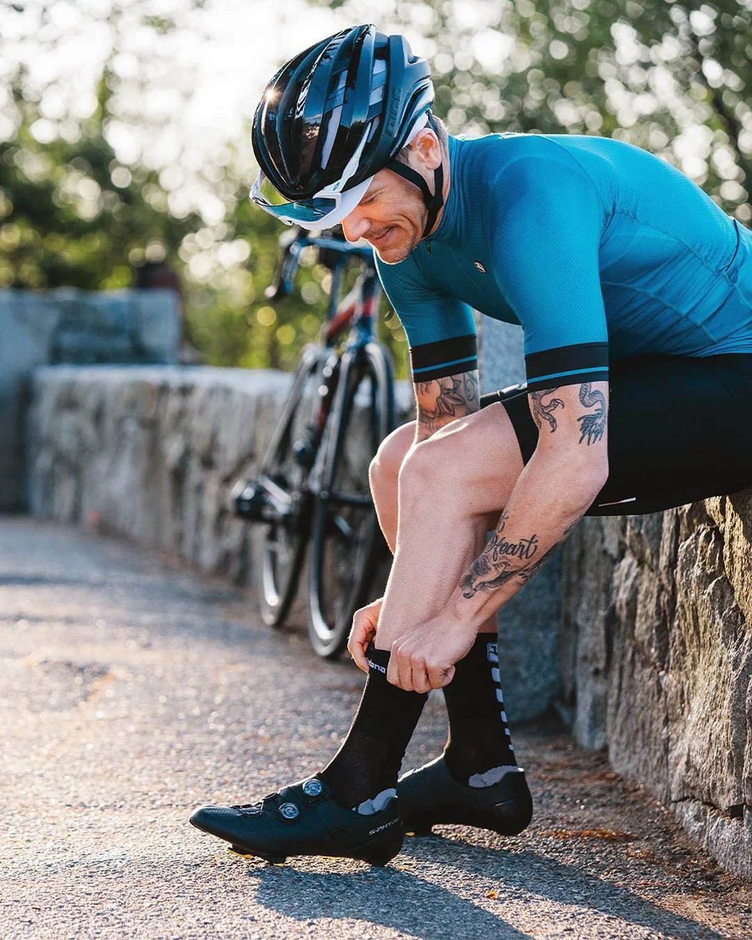 Ein Radfahrer mit Helmm, blauer Radtrikot und Tattoos sitzt auf einem Steinmauer und zieht seine Fahrradschuhe an, mit einem Fahrrad im Hintergrund, unter Sonnenlicht und Bäumen.