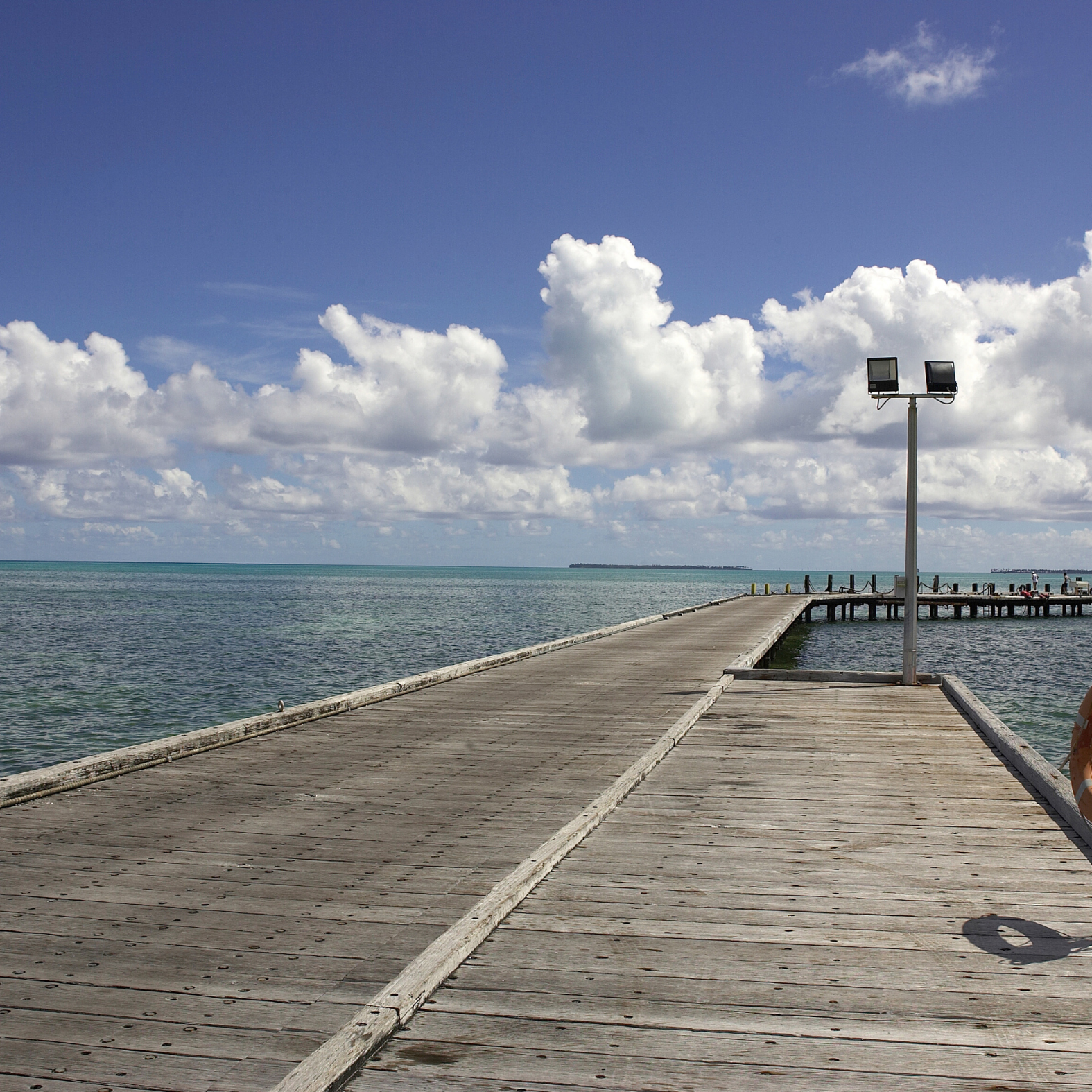 A wooden pier extends into the ocean with a cloudy sky overhead, and a lamp post on the right side of the pier.