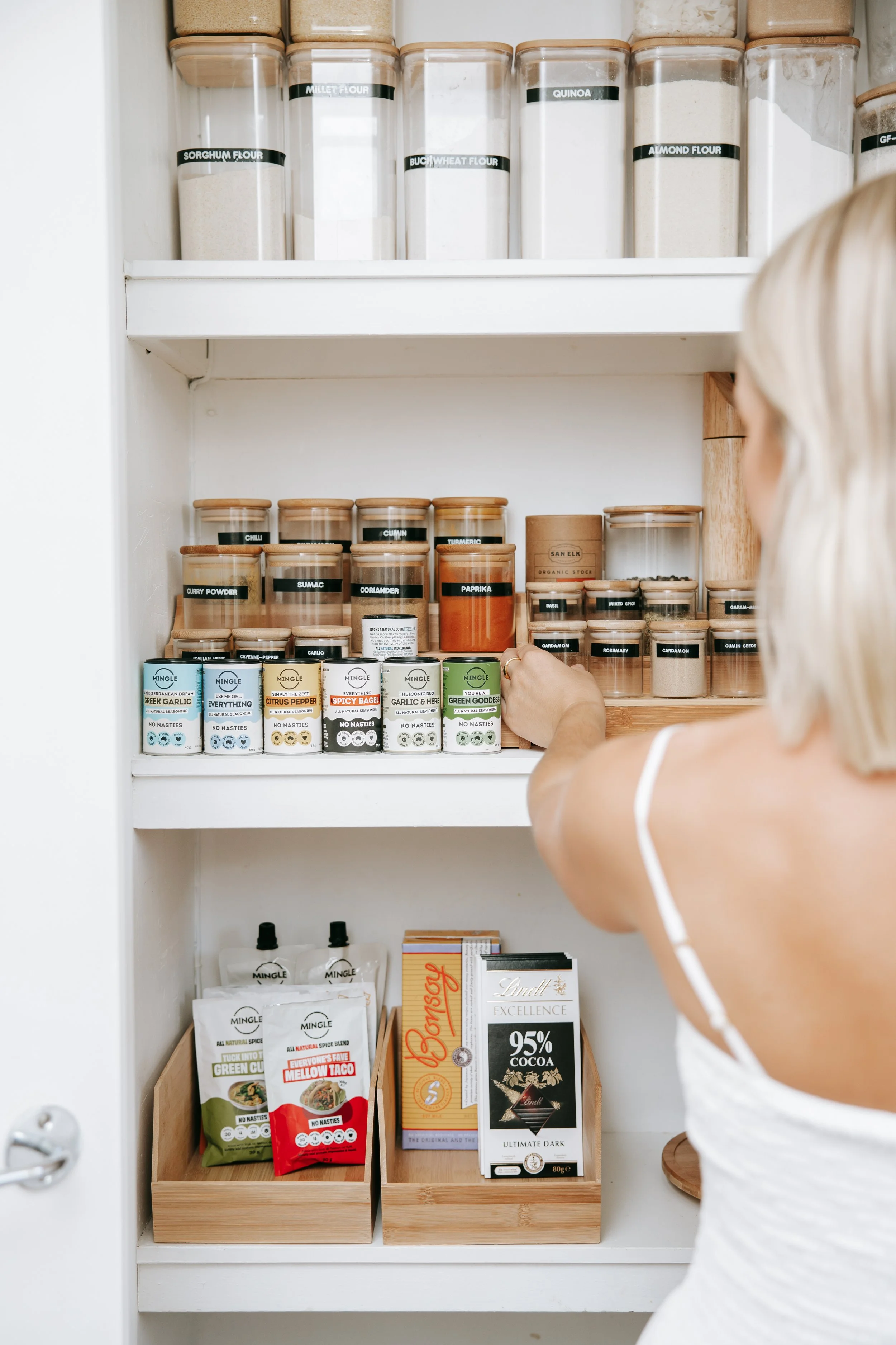 A person reaching for spice jars in a pantry with jars labeled for flour, quinoa, and spices, and boxes of chocolate and snack pouches on the bottom shelf.