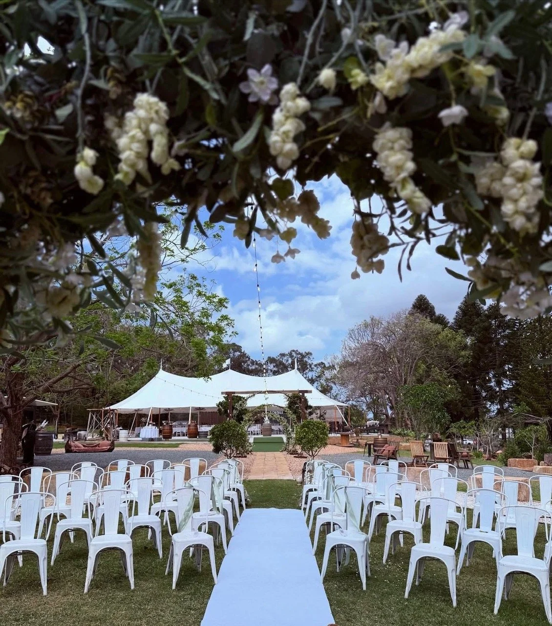 Outdoor wedding ceremony setup with white chairs, an aisle, a tented area in the background, and decorative greenery and flowers.