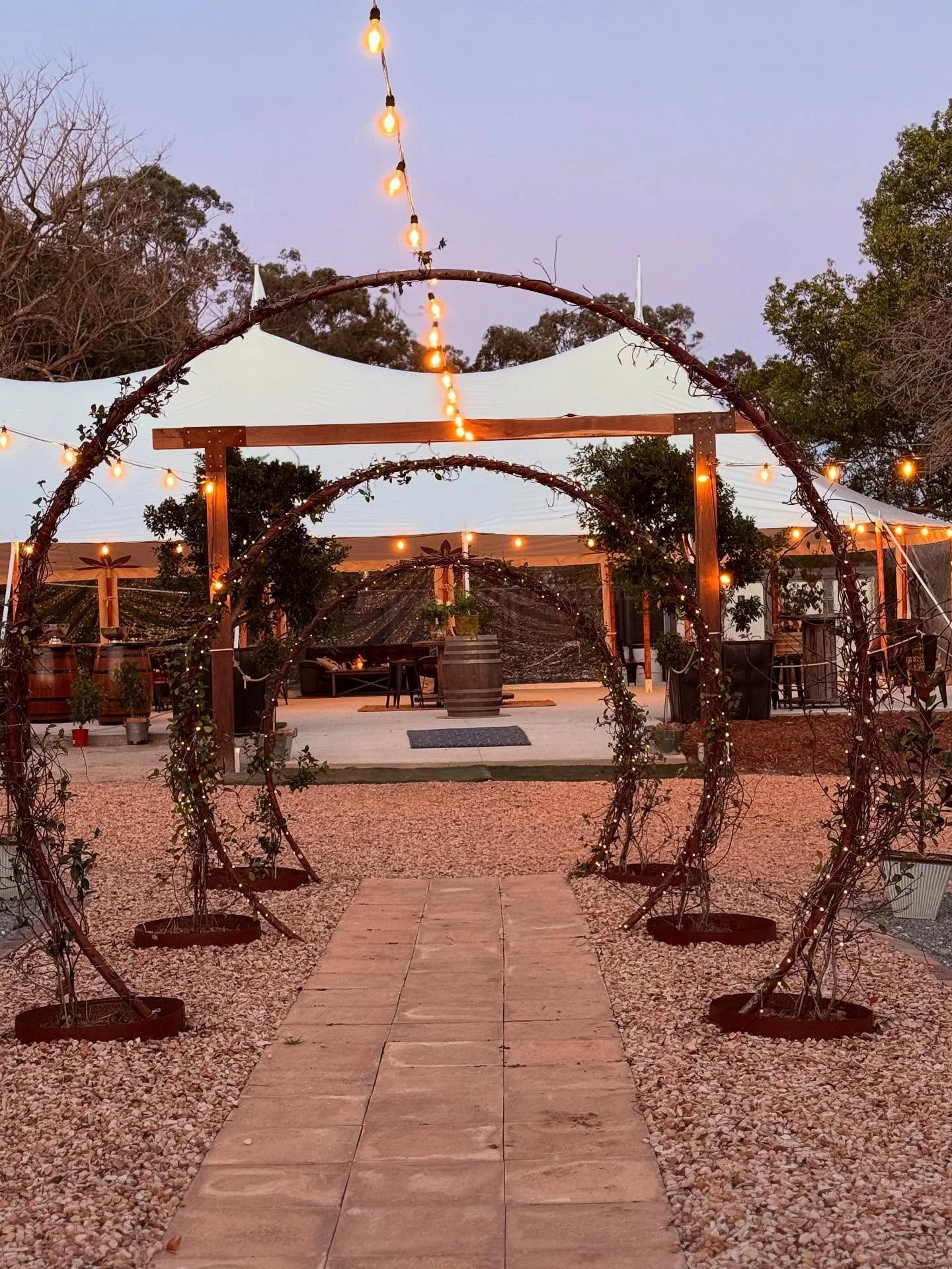 An outdoor event venue with a pathway leading to a raised wooden floor under a white canopy, decorated with string lights and circular wooden arches wrapped with vines and small lights, surrounded by trees.