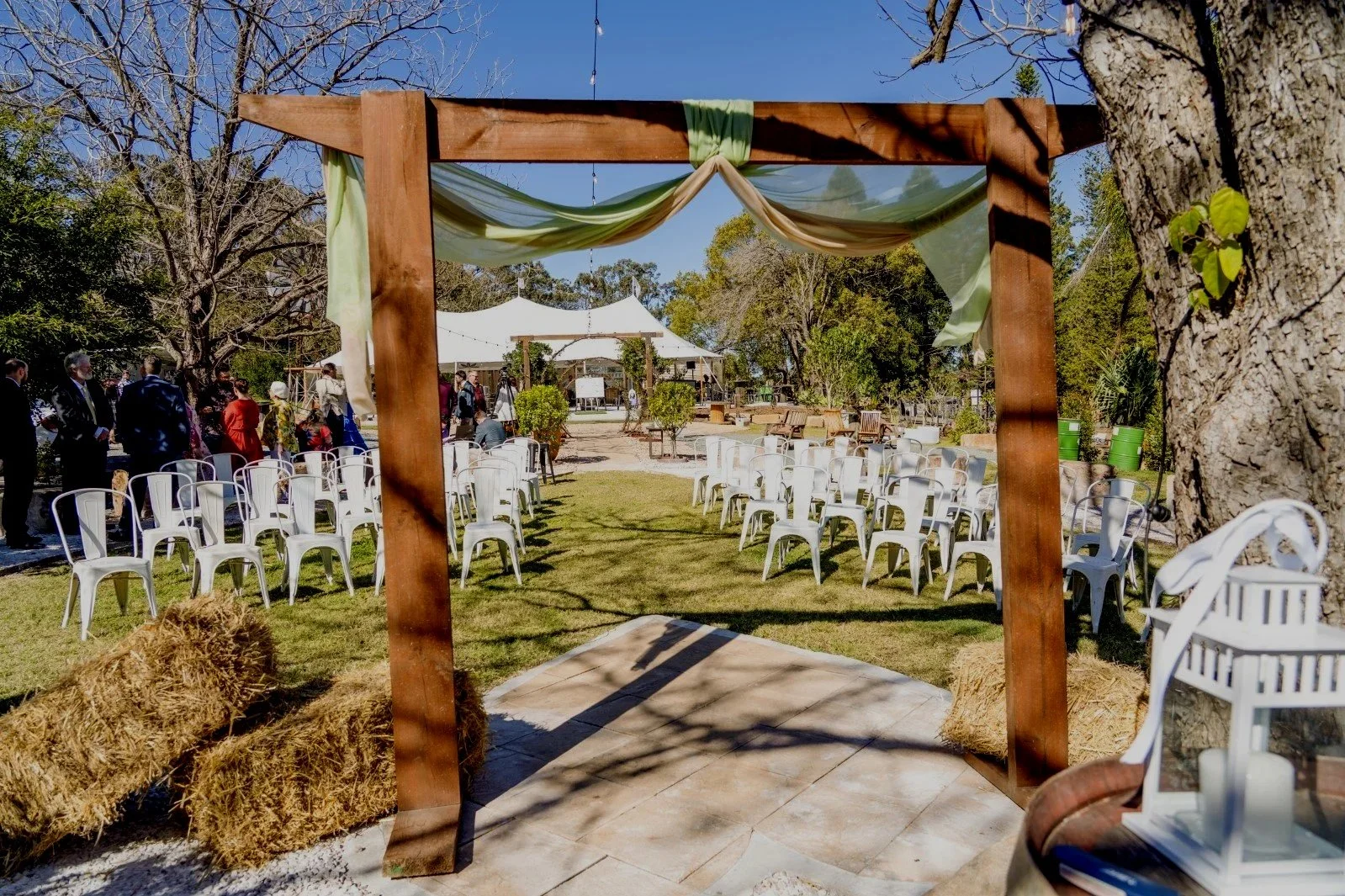 Outdoor wedding ceremony setup with white chairs arranged on grass, a wooden arch with draped fabric, hay bales, and a tent in the background under a clear blue sky.