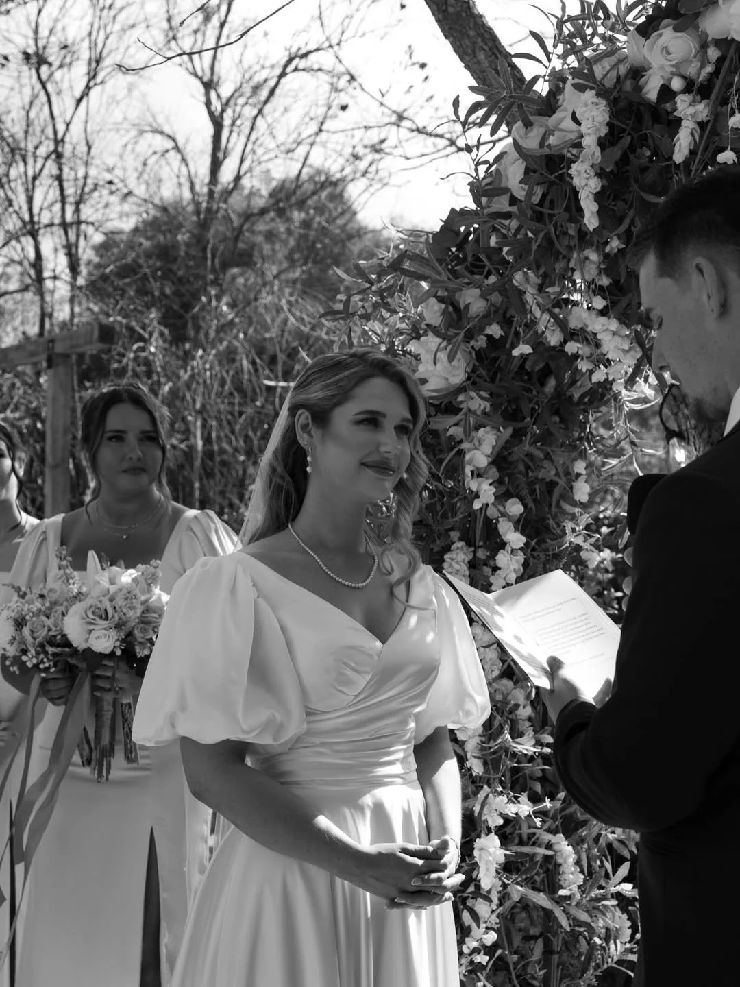 A black-and-white photo of a wedding ceremony outdoors, showcasing a bride in a white dress with puffed sleeves and pearls, standing in front of a floral backdrop, exchanging vows with a groom holding a paper, with two bridesmaids in the background, one holding a bouquet.