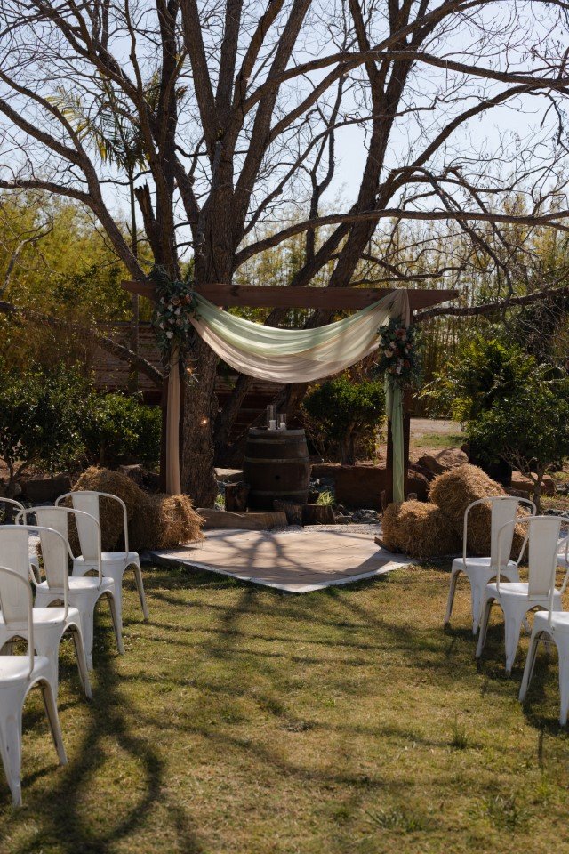 An outdoor wedding setup with a wooden arch decorated with fabric and flowers, facing a small stage with chairs arranged on both sides, surrounded by trees and bushes.