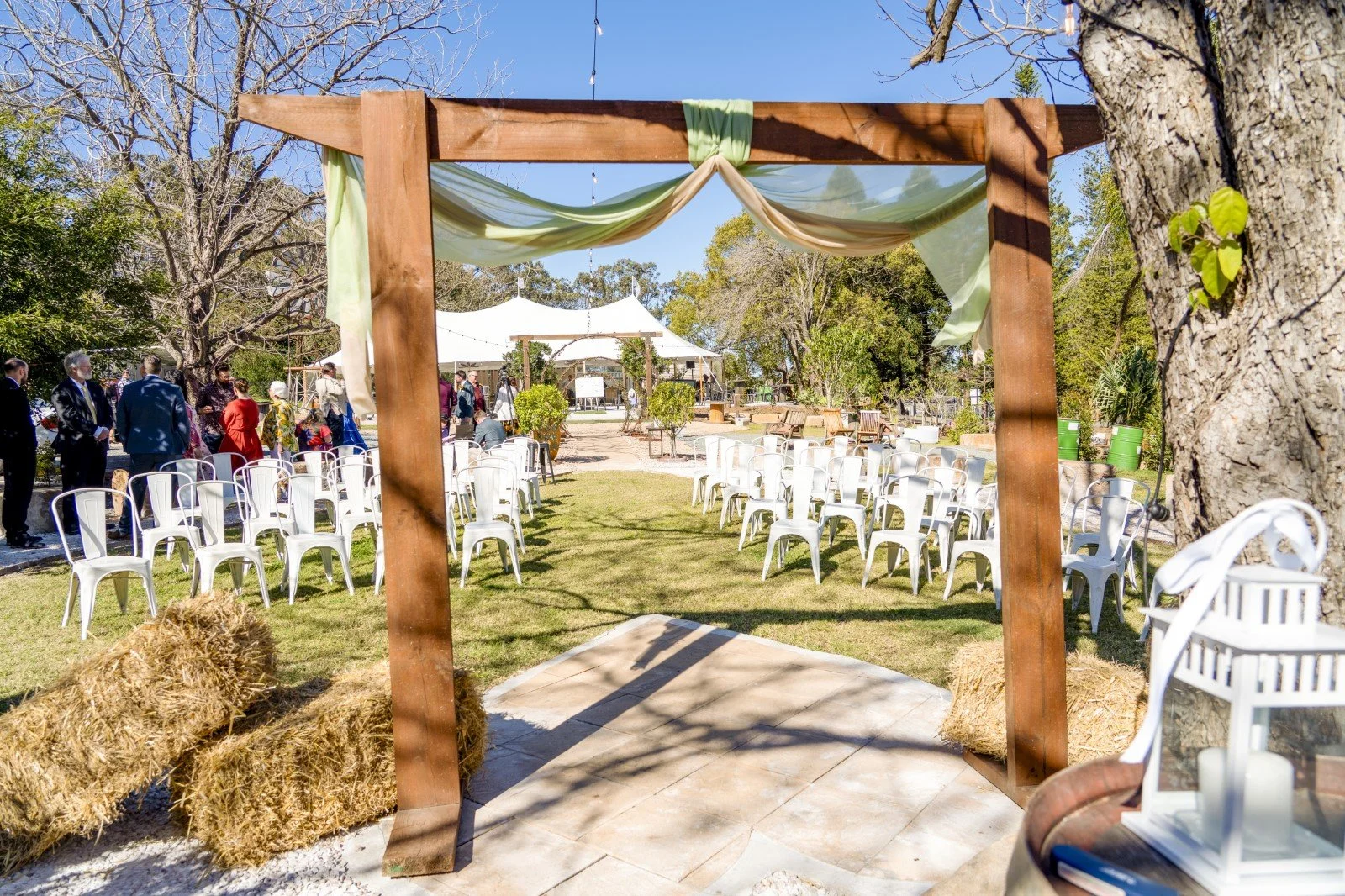 Outdoor wedding ceremony setup with white chairs arranged in rows facing a small altar, under a wooden arch decorated with green and beige drapes, surrounded by hay bales and trees on a sunny day.