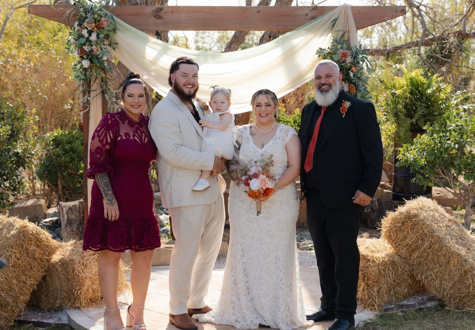 Group of six people at an outdoor wedding, standing in front of a wooden arch decorated with flowers and draped fabric, with hay bales on either side, during daytime.