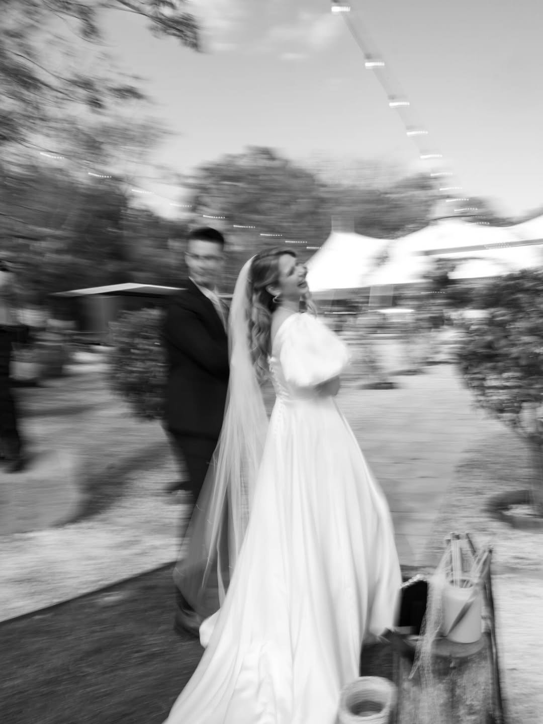 Black and white photo of a bride and groom at their wedding, walking outdoors, with a blurred background and string lights overhead.