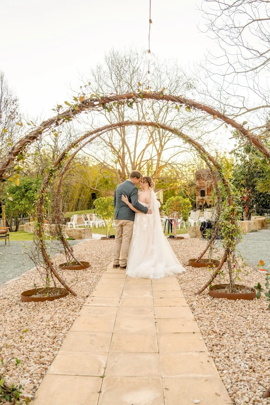 A bride and groom sharing a kiss under a rustic arch at an outdoor wedding ceremony during sunset, surrounded by trees and decorative elements.