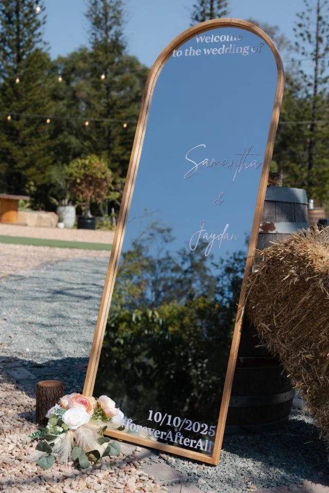 An outdoor wedding sign with gold frame that reads "Welcome to the wedding of Samantha & Jaydan, 10/10/2025, Forever and Always". The sign is decorated with white and pink flowers at the base and is reflected in a mirror surrounded by trees and strin
