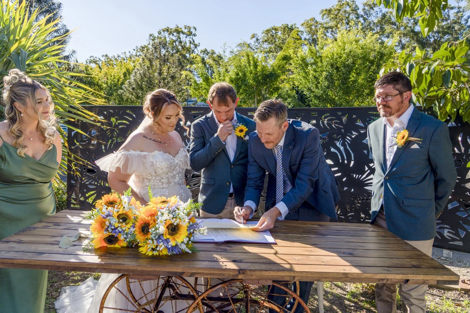 A wedding ceremony outdoors with six people gathered around a wooden table, signing a document. There are two women, two men, and a flower arrangement of sunflowers and other flowers on the table. The background shows green trees and blue sky.