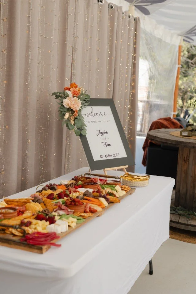 A cheese and charcuterie platter on a white table with a wedding welcome sign in the background decorated with flowers and string lights.