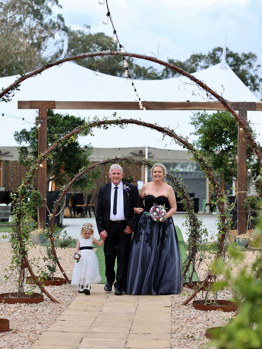 A wedding ceremony outdoors with a bride, groom, and a young girl walking down a wooden path under decorative arches with greenery. The bride is wearing a black strapless gown and holding a bouquet, while the groom and girl are dressed in formal atti
