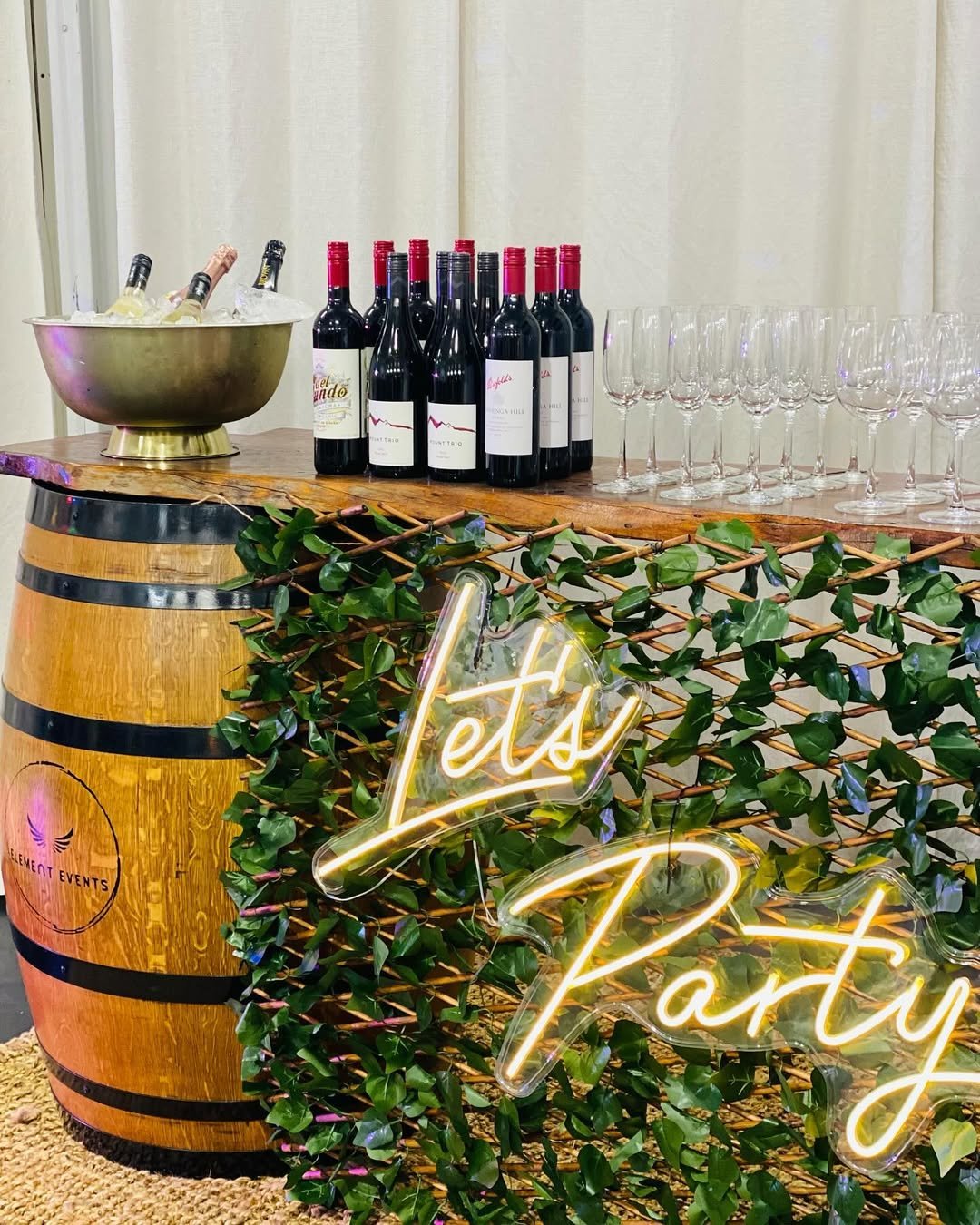Wine bottles, glasses, ice bucket, and greenery on a wooden barrel with a neon sign that reads 'Let's Party' at a celebration event.