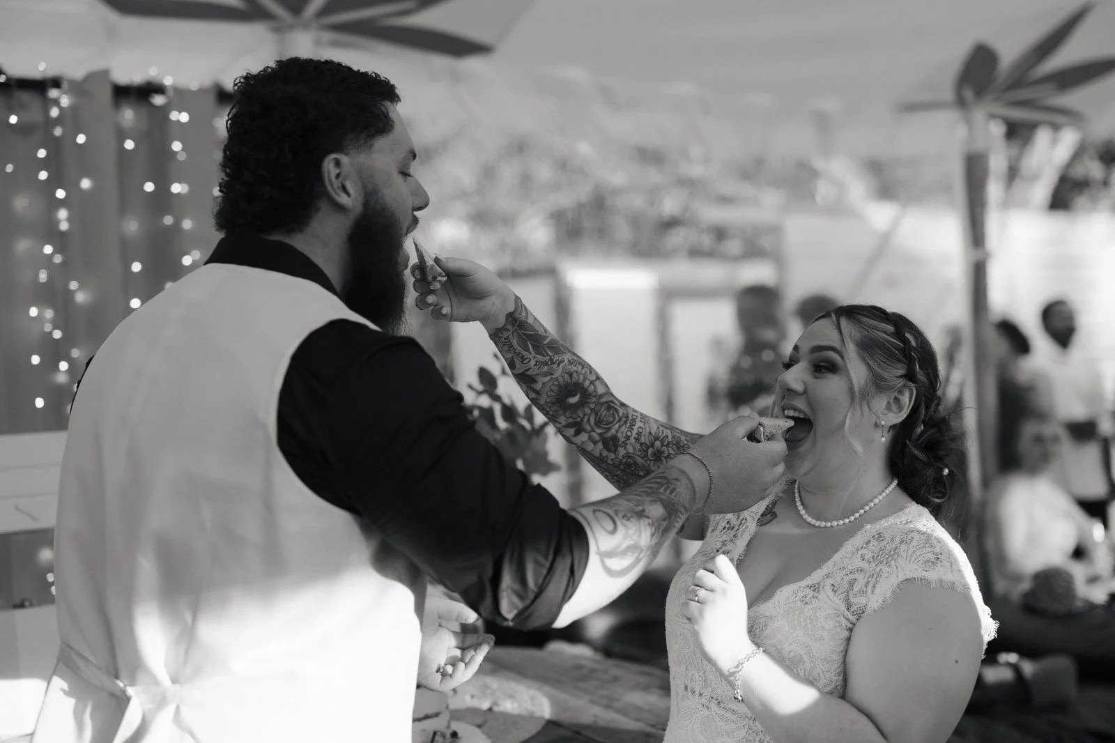 A man with tattoos and a beard offers cake to a woman in a lace dress during a celebration, possibly a wedding, with people and decorations blurred in the background.
