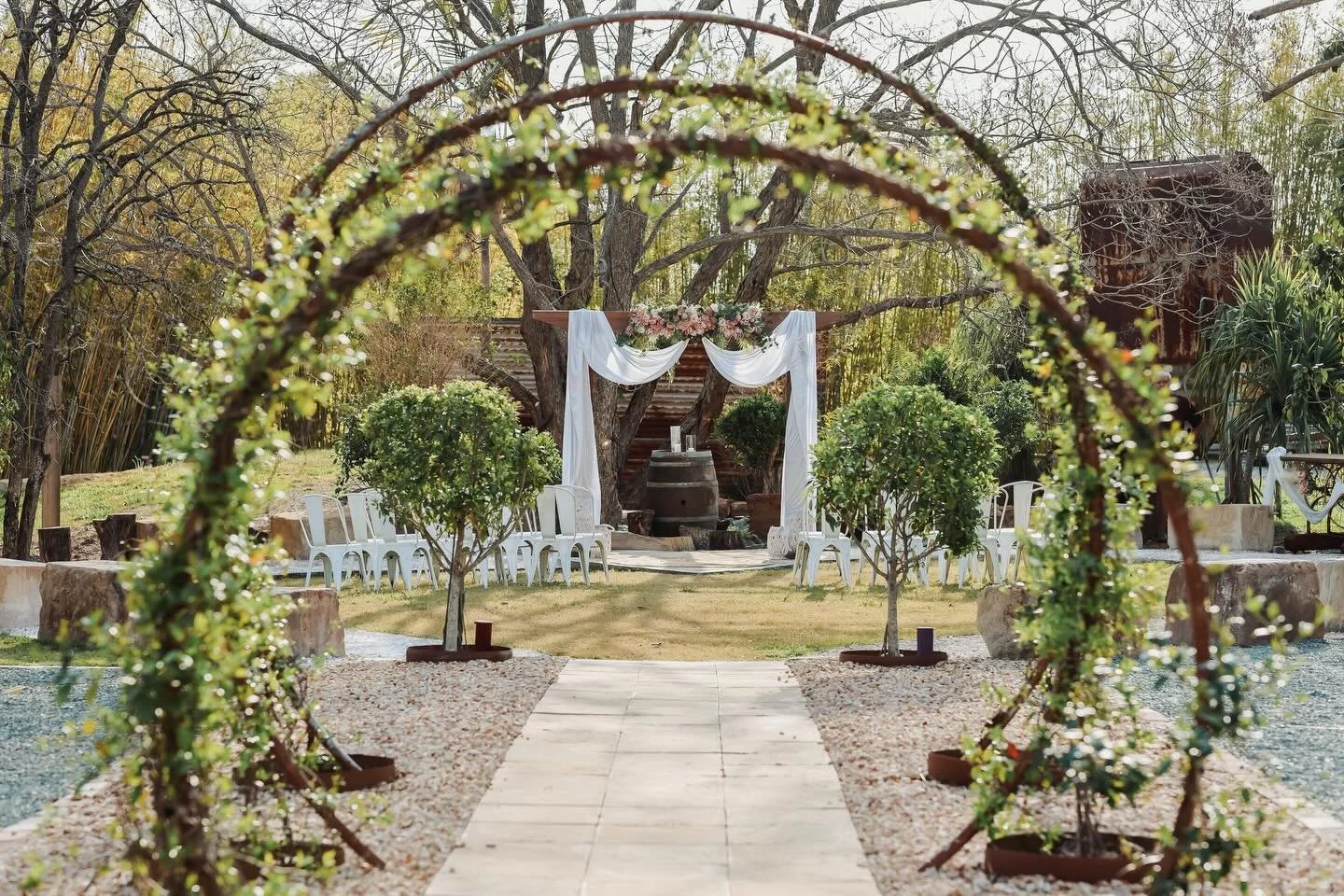 Outdoor wedding setup with a wooden arch decorated with white drapes and flowers, leading to a ceremony area with white chairs and small trees, set amidst greenery and trees.