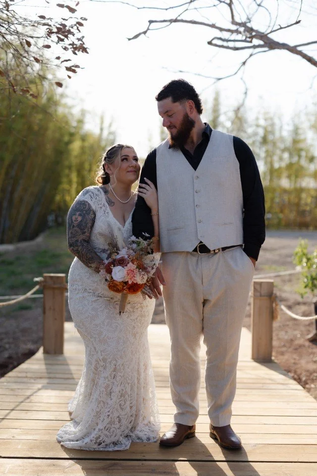 Bride holding a bouquet of flowers, standing next to groom on a wooden bridge outdoors, with trees in the background during daytime.