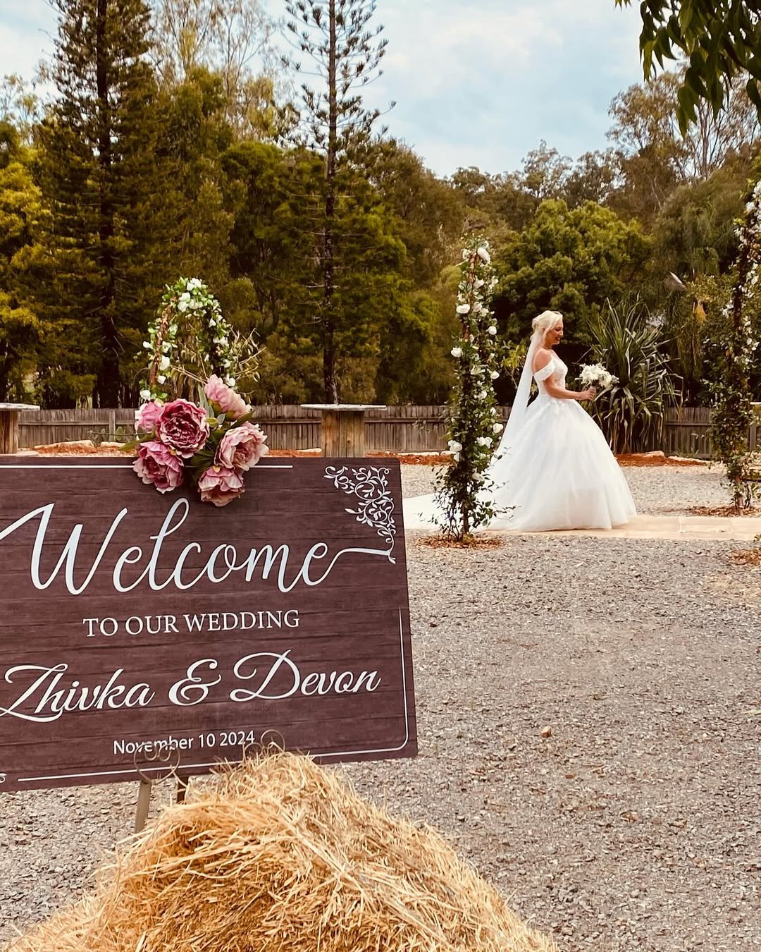 A bride in a white wedding dress holding a bouquet stands outdoors near floral arrangements and a welcome sign that reads, 'Welcome to our wedding Zhuka & Devon, November 10, 2024.' The setting is natural with trees and a wooden fence in the backgrou