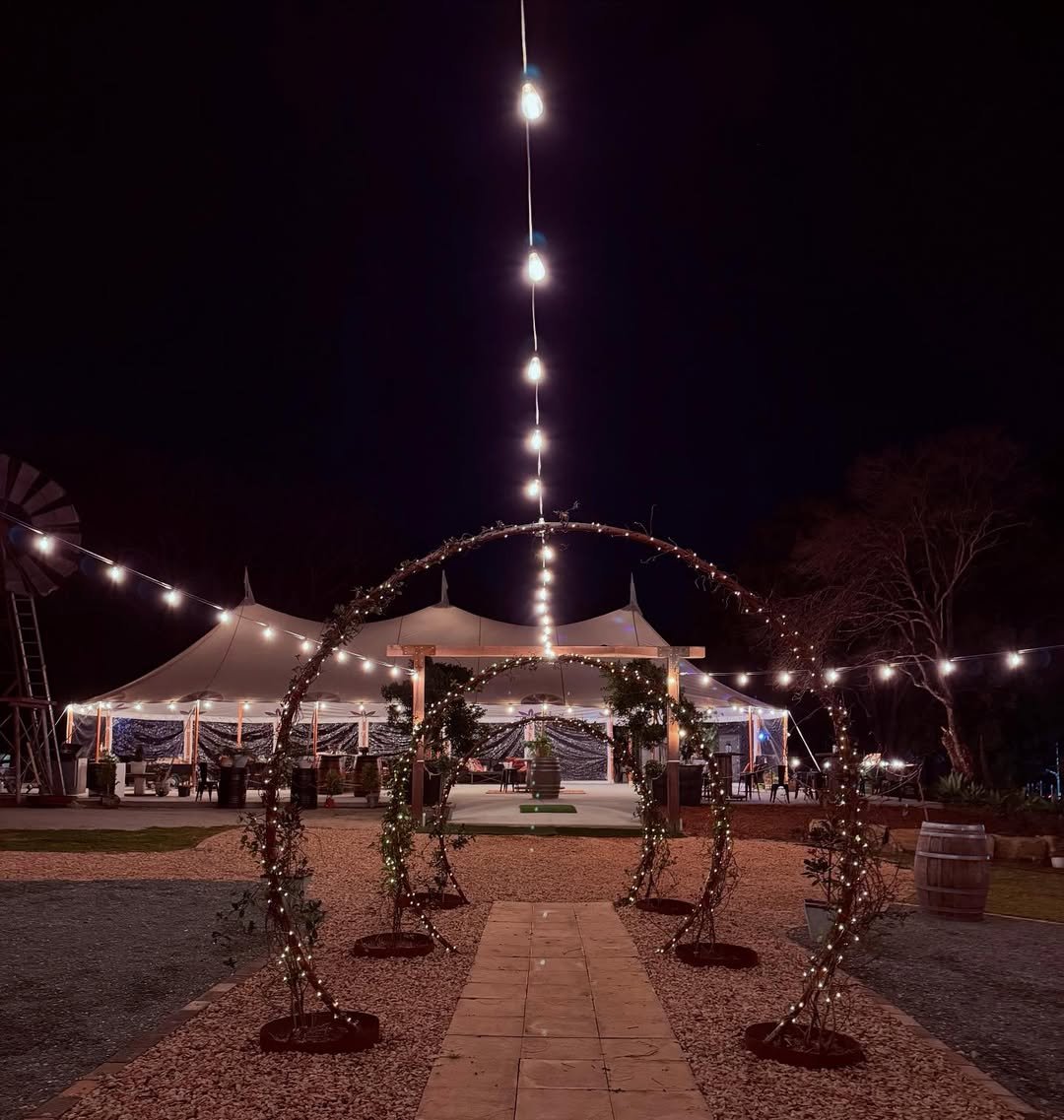 Night scene of an outdoor event space decorated with string lights, a tent in the background, and a circular archway with fairy lights in the foreground. There is a gravel path leading to a stage area.