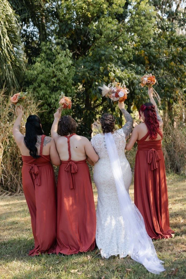 Bride with three bridesmaids outdoors, all holding bouquets and raising their arms.