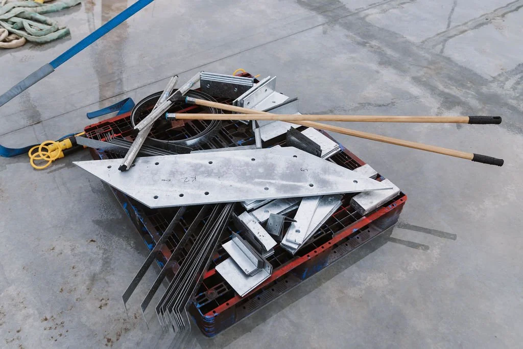 Metal construction tools and parts, including metal beams, blades, and sticks, resting on a plastic pallet on a concrete floor at a construction site.