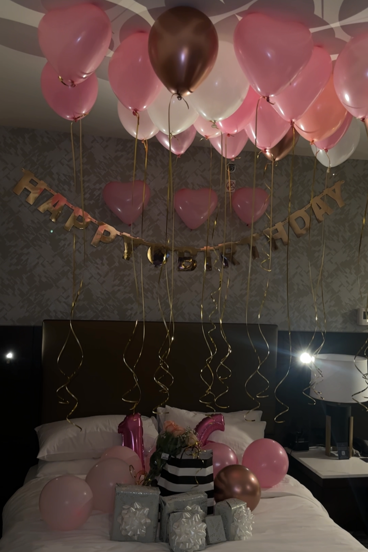 Decorated hotel room with pink and bronze balloons, a birthday banner, gifts, and a birthday cake on the bed.
