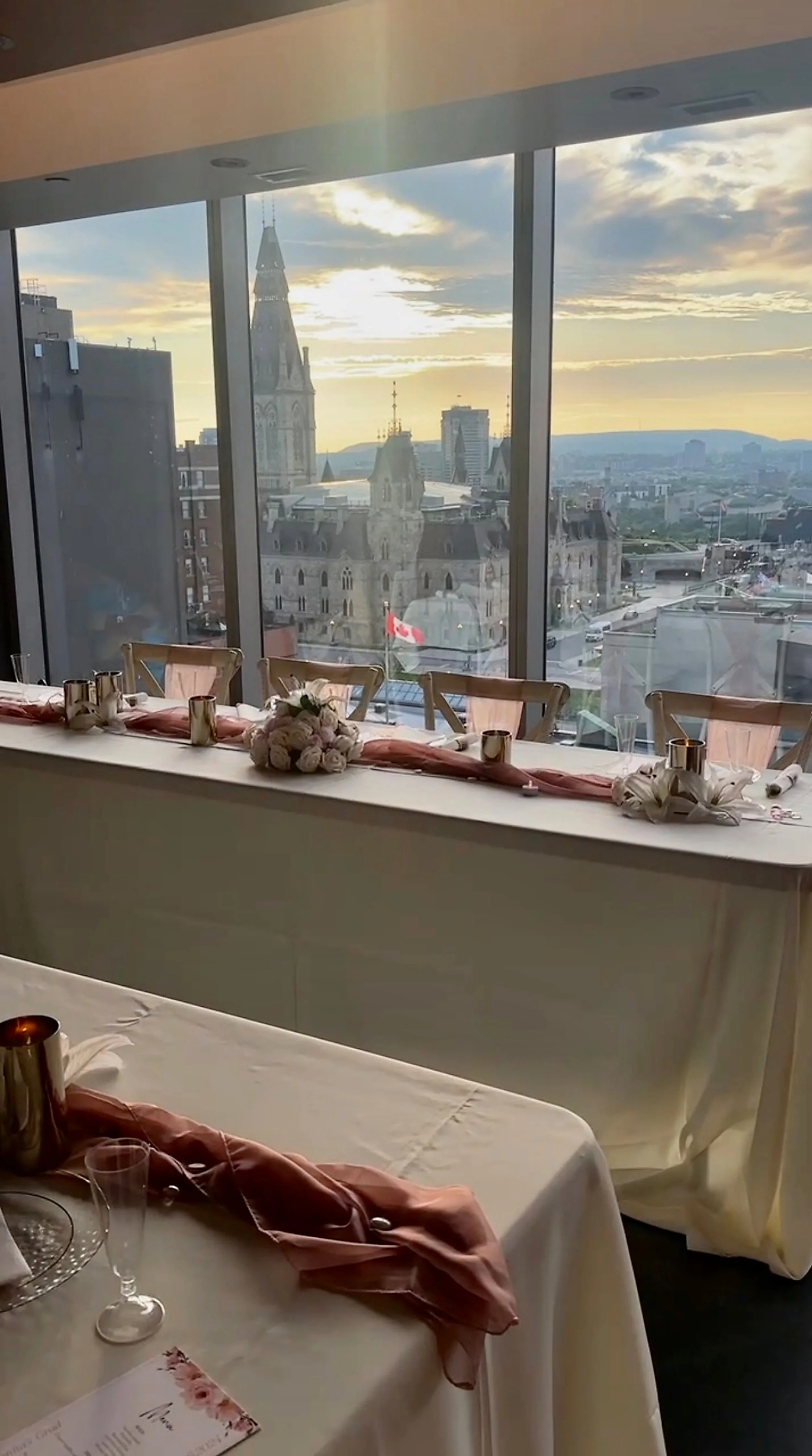 Interior view of a decorated dining table with pink and white accents, set for an event, overlooking a city skyline with historic buildings and a church spire during sunset.