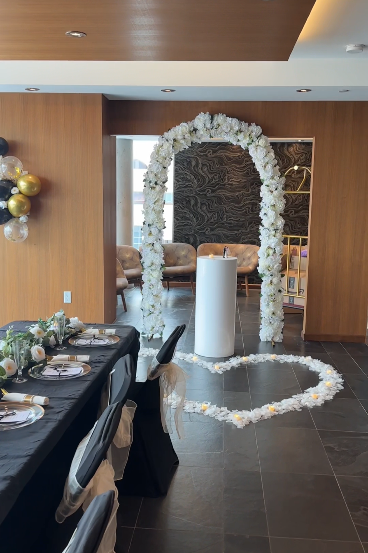 A decorated event space with a white floral arch, heart-shaped flower petal arrangement on the floor, candles, and a table set for a celebration or wedding.