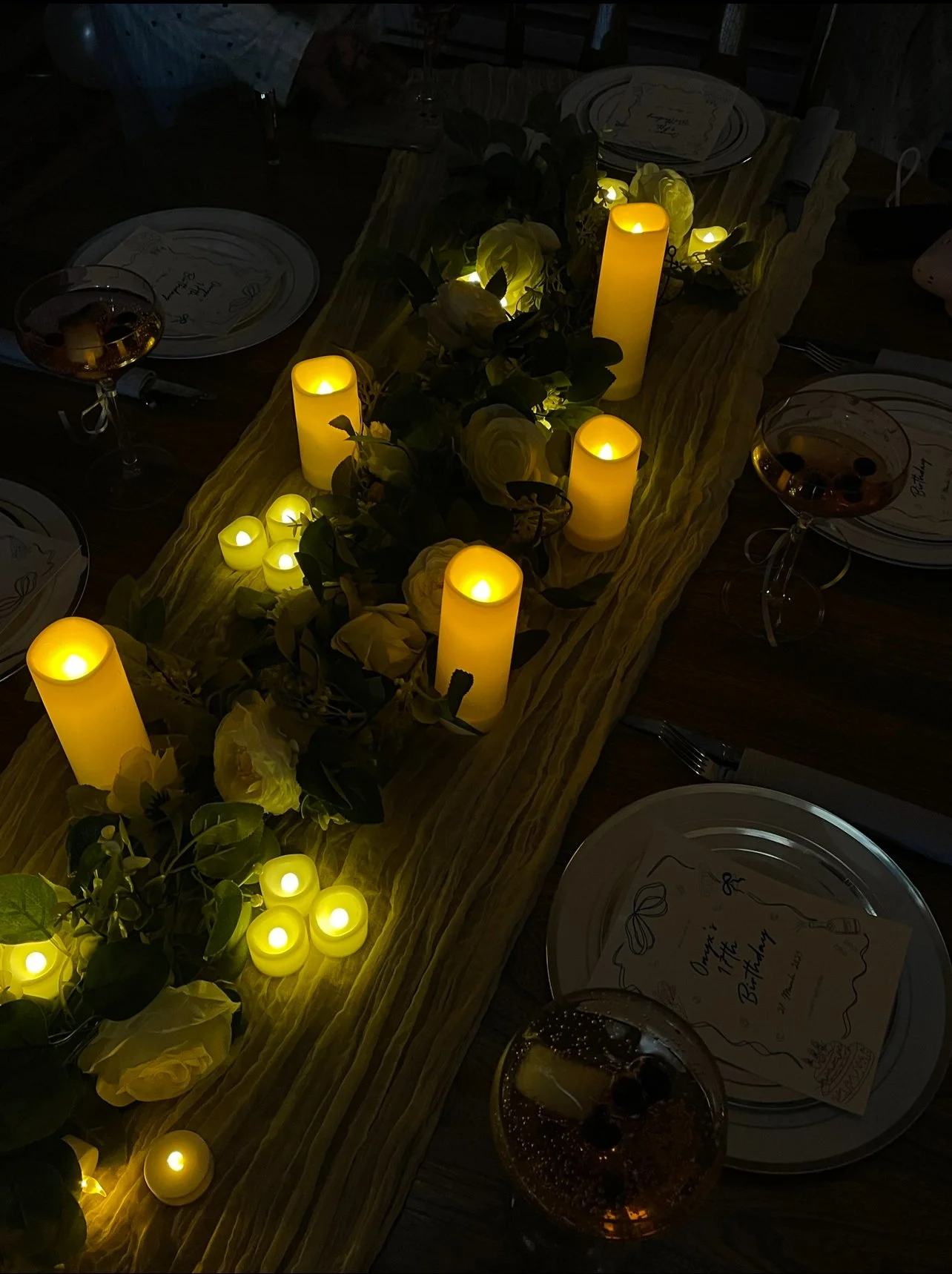 A dinner table decorated with a yellow table runner, yellow candles, and white flowers, illuminated by candlelight, with plates and glasses set at each place.