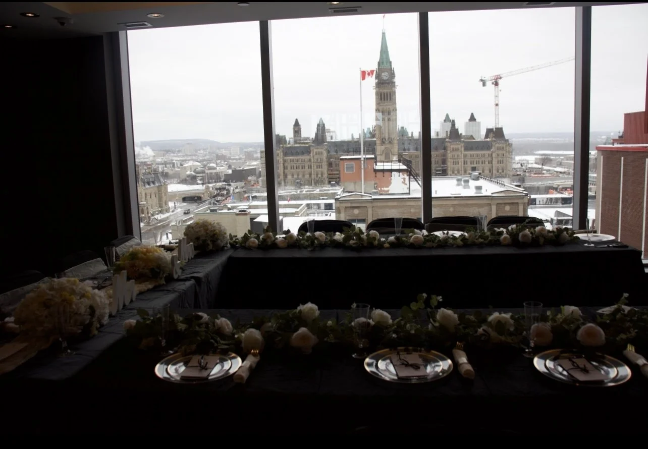 Elegant dining table decorated with flowers and napkins in front of large windows showing a city skyline with historic building and clock tower.