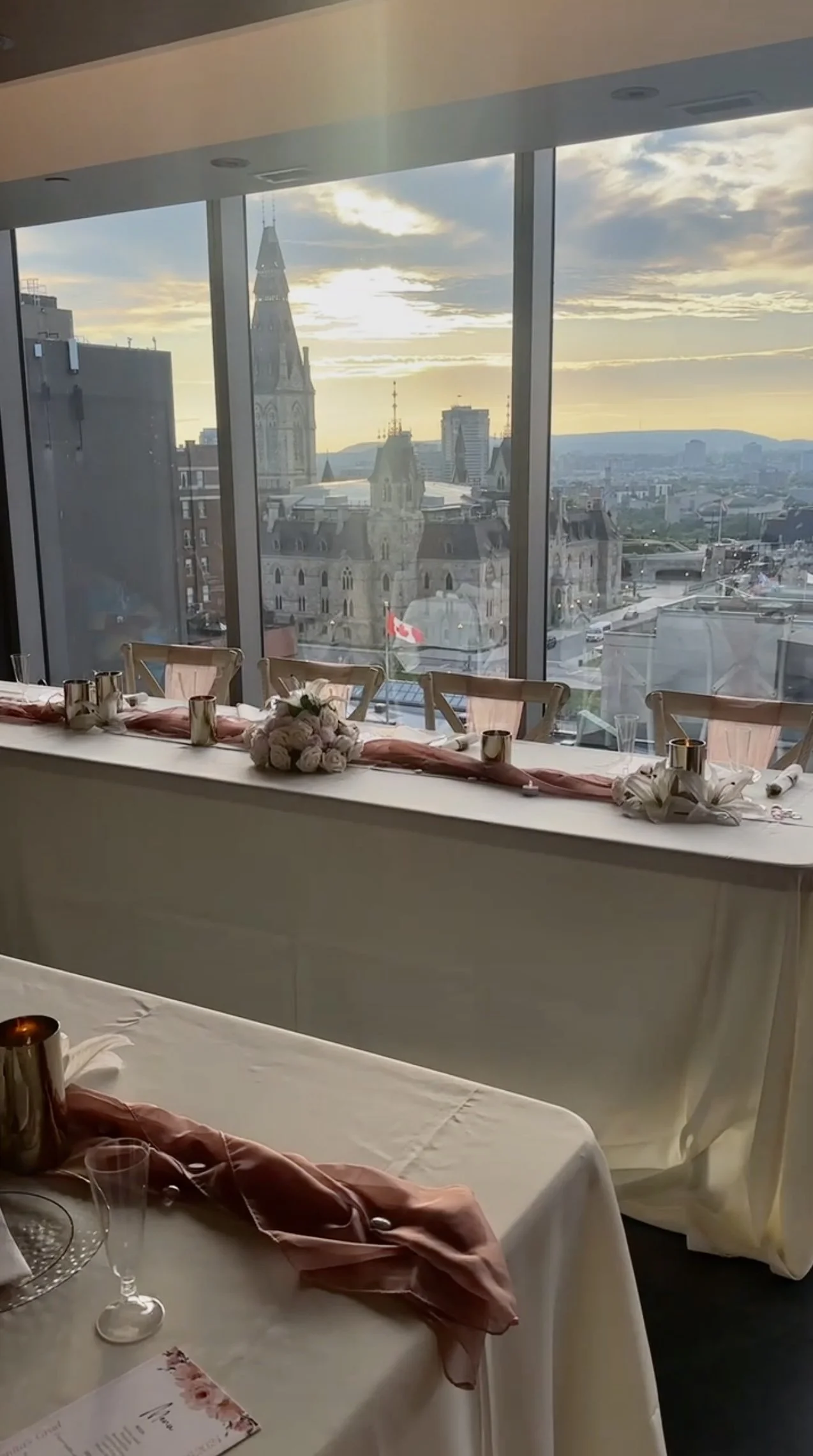 Set up for a celebration or wedding reception with decorated tables, napkins, and floral centerpieces, overlooking a cityscape with church spires and a Canadian flag in the background.
