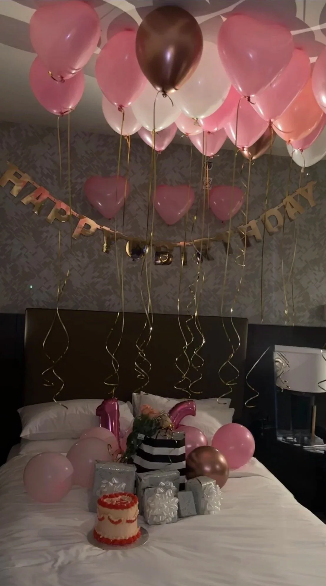 Decorations for a birthday celebration on a bed, including pink, white, and rose gold balloons, a birthday cake, wrapped presents, and a banner that reads 'Happy Birthday.'