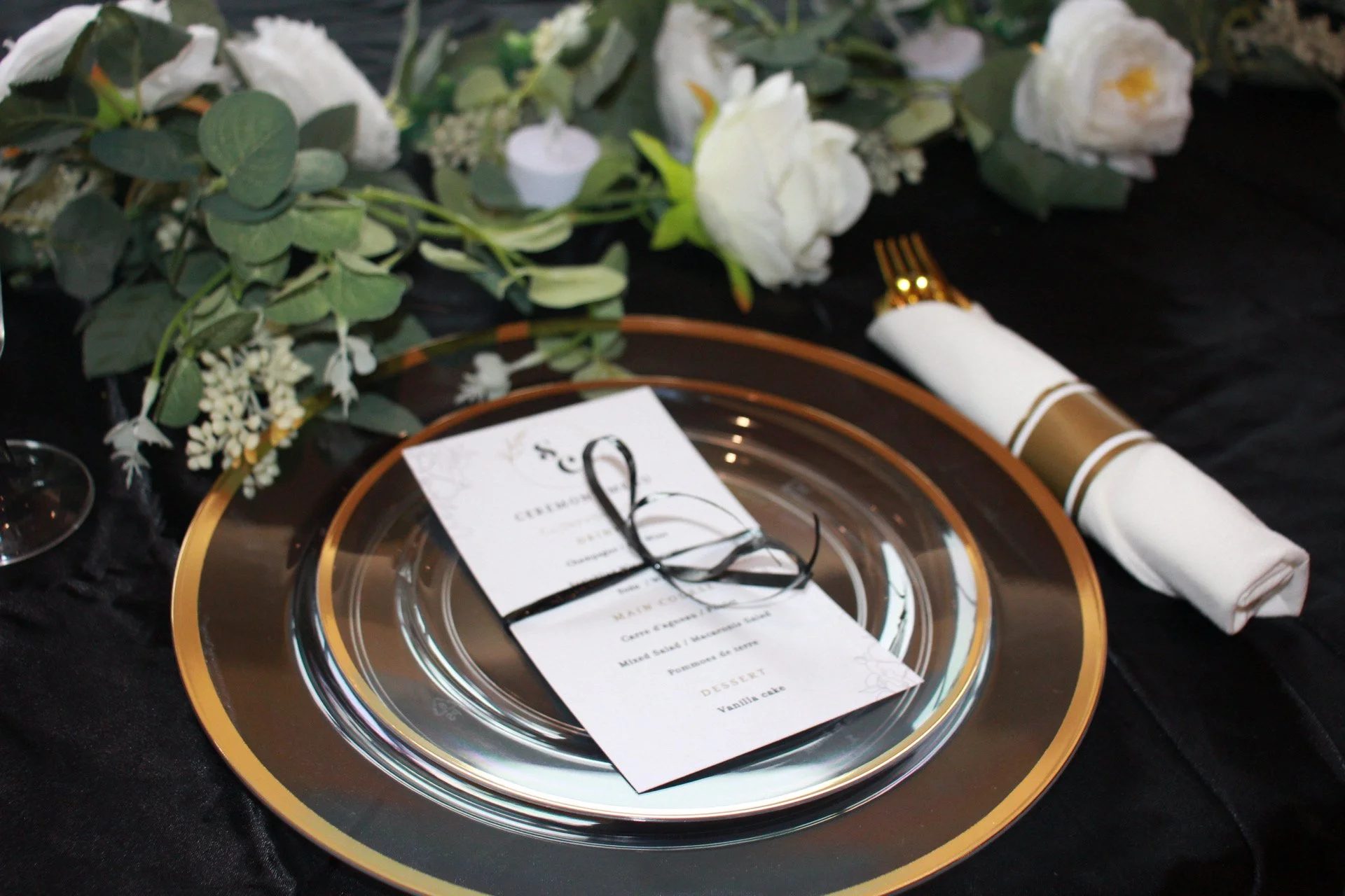 Elegant place setting with a gold-rimmed glass plate, a black and white menu tied with black string, a white napkin with gold accents, and a floral centerpiece with white roses and greenery.