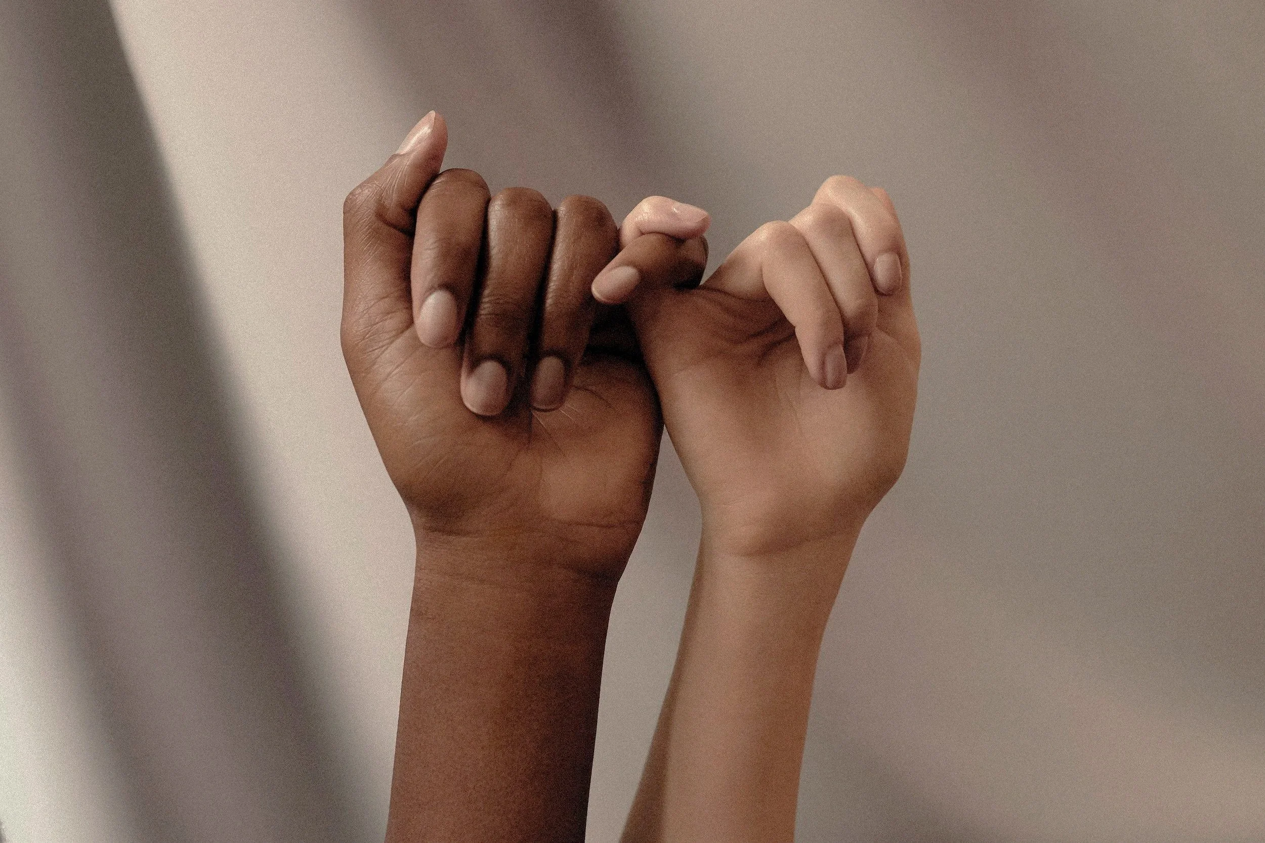 Two hands, one dark-skinned and the other light-skinned, holding each other's pinky fingers.