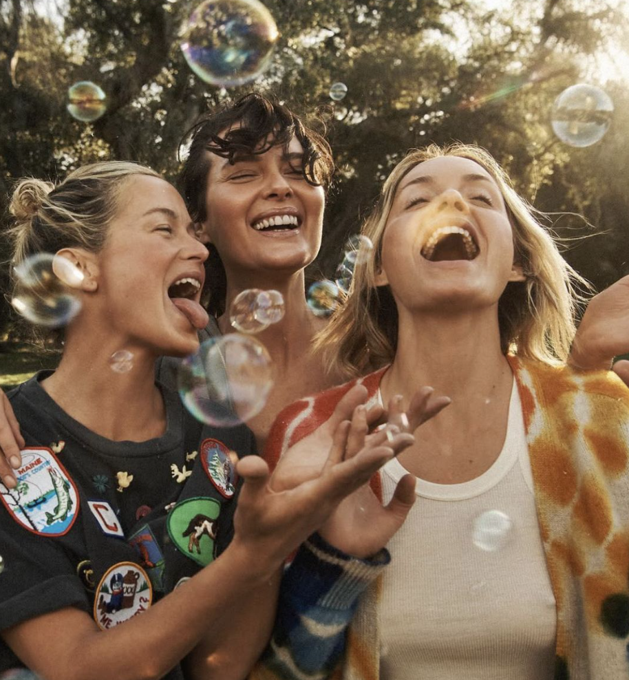 Three women smiling and laughing outdoors with soap bubbles floating around them, sunlight filtering through trees in the background.
