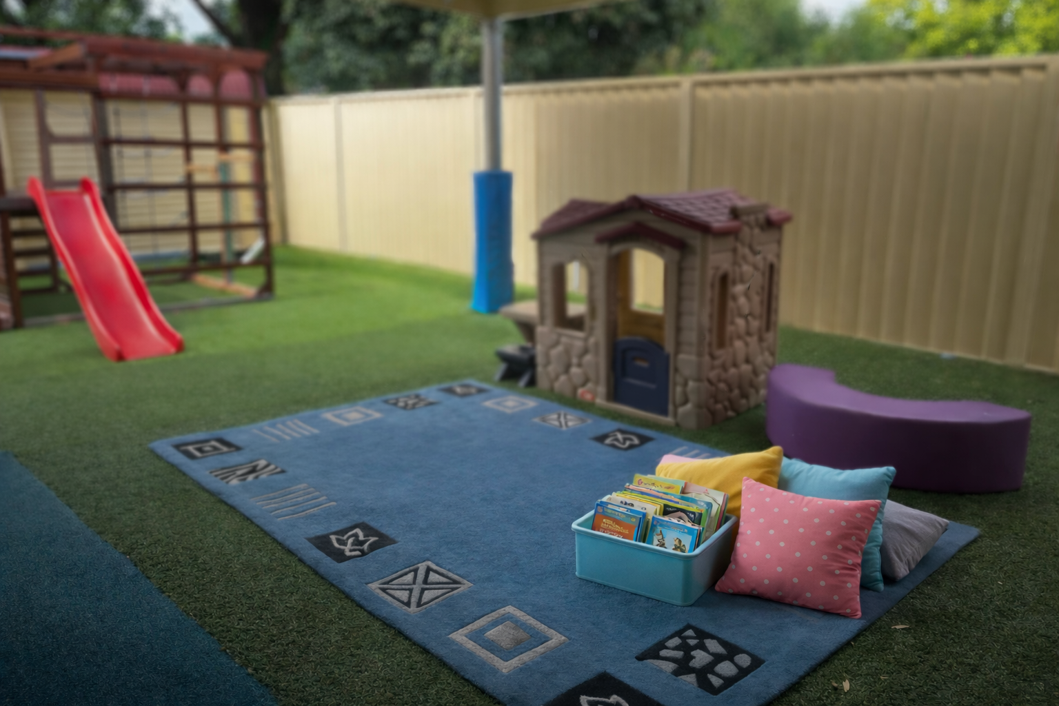A backyard play area with a blue rug, colorful pillows, a basket of books, a small plastic house, and a purple seating area, enclosed by a wooden fence with a slide and a play structure in the background.