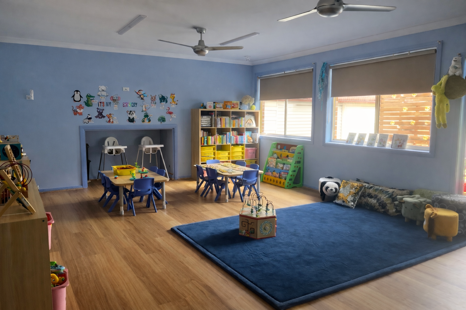Children's playroom with blue walls, wooden floor, and a large blue rug, featuring small tables and chairs, a bookshelf filled with books, assorted toys, stuffed animals, and colorful wall decorations.