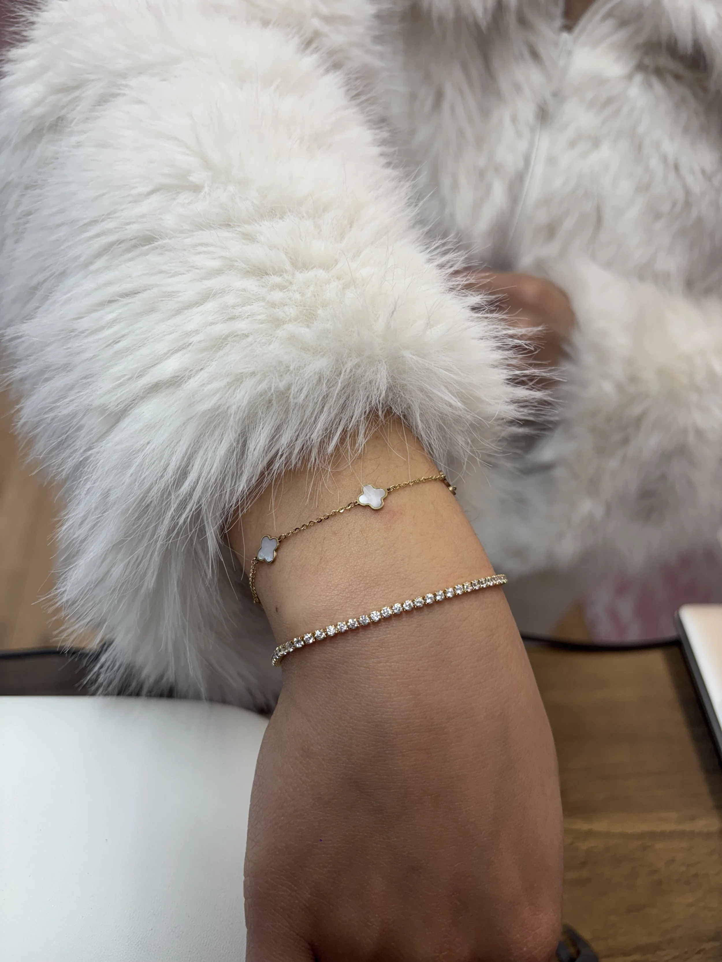 A person's wrist with two bracelets, one delicate gold chain with white inlaid stones and a second gold chain with small sparkling diamonds. The person is wearing a white furry long-sleeve garment.