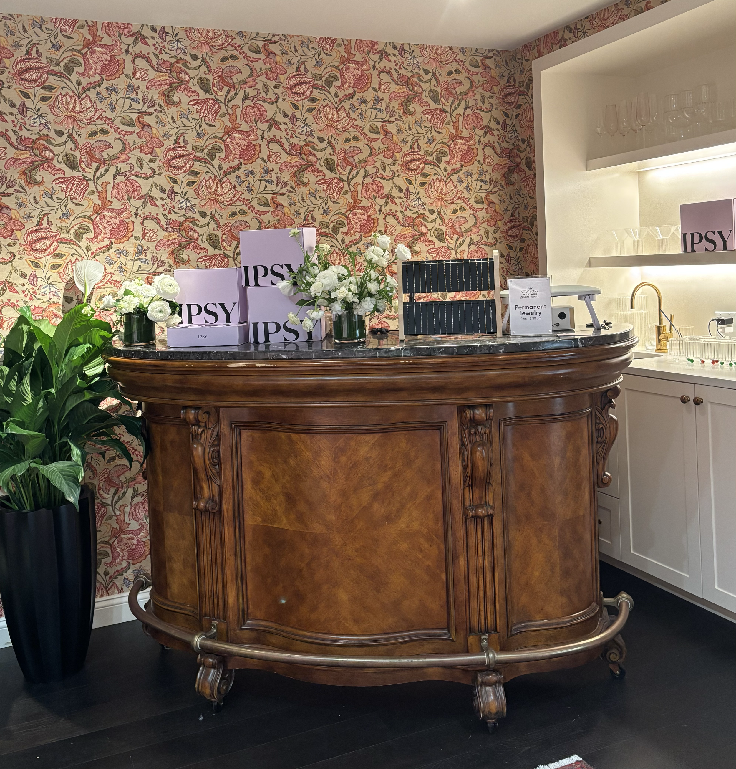 An elegant wooden bar counter with a marble top in a room with floral-patterned wallpaper. The bar has brass feet and details, and there are pink boxes with the word 'IPSY', a bouquet of white flowers, and shelves with glassware and a pink sign that 
